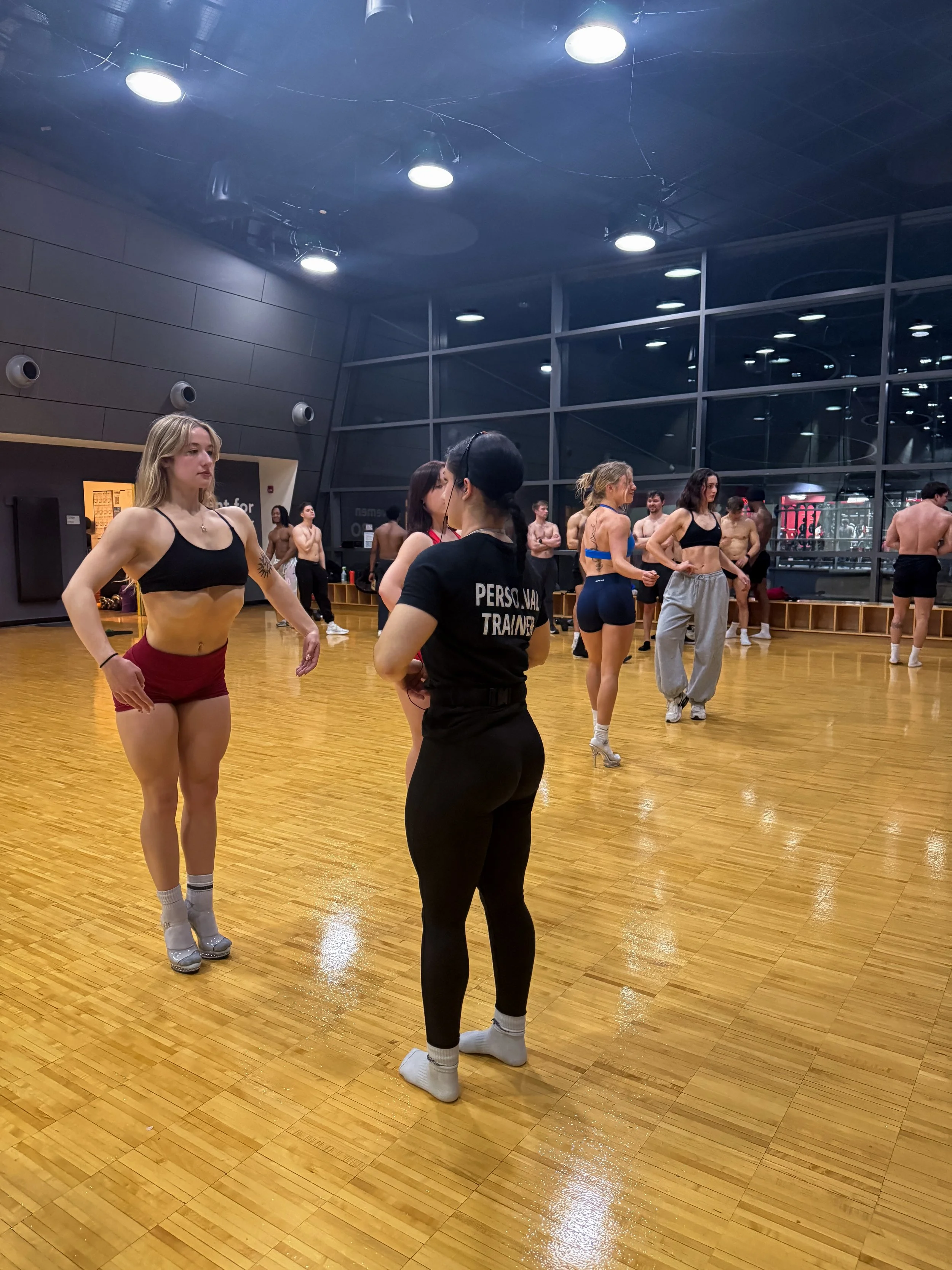 A group of people in a dance or fitness class in a gym or dance studio with large windows and wooden floors. The women are dressed in workout attire, and some men are shirtless. The instructor appears to be demonstrating or talking to the class.