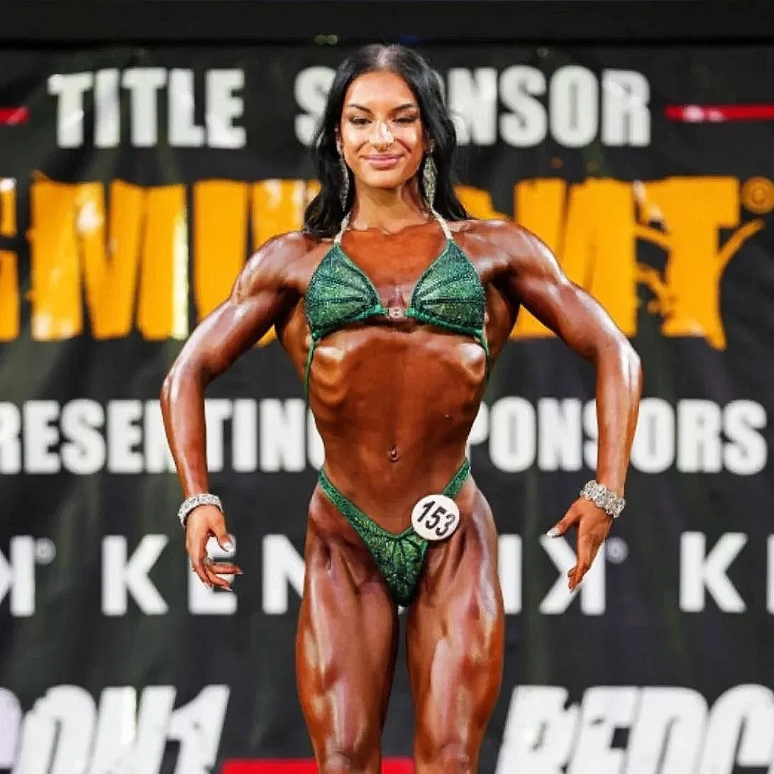 A female bodybuilder on stage in a green bikini, smiling with arms flexed, showcasing muscular physique during a competition.