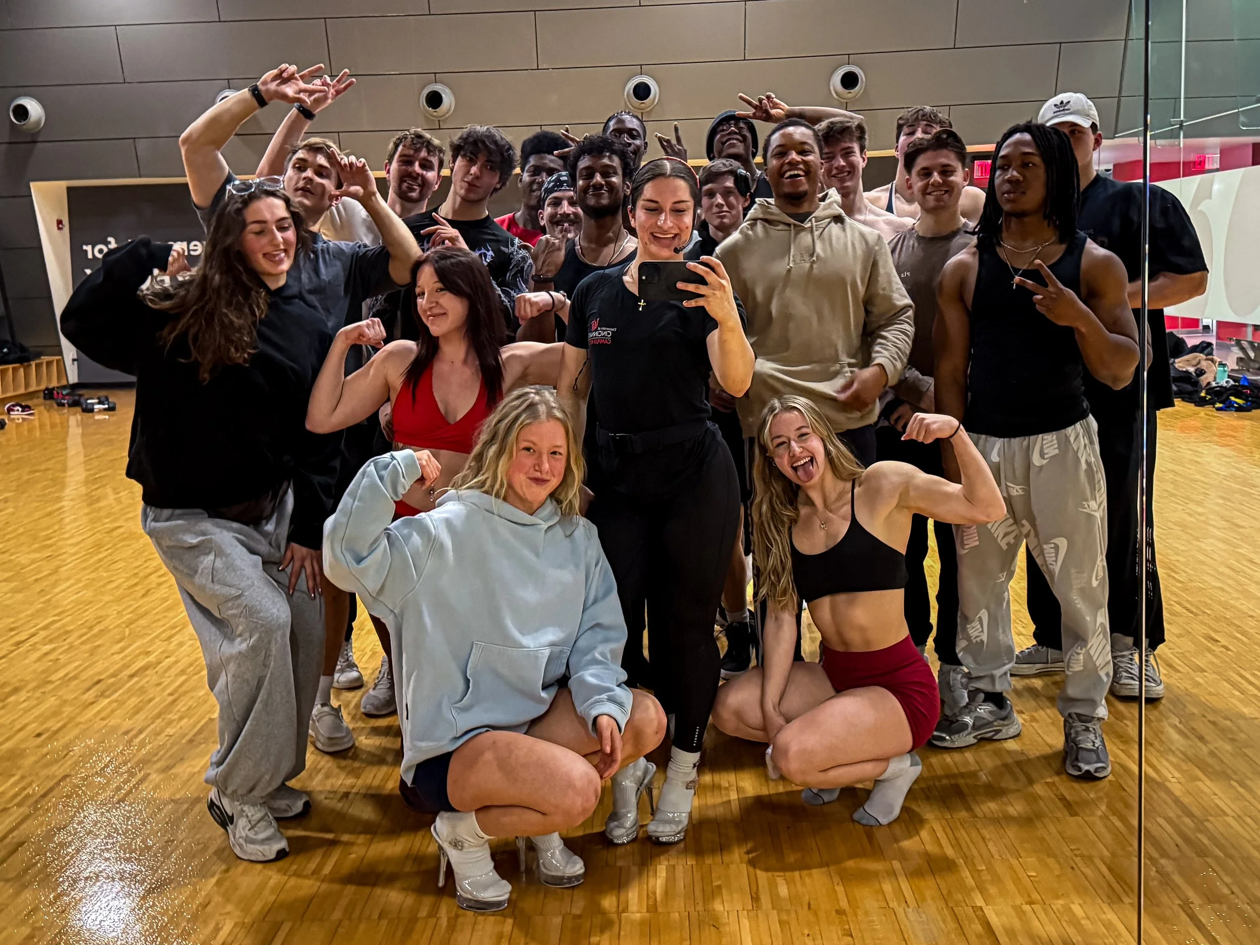 A group of young adults in athletic clothing taking a group selfie in a gymnasium. Some are smiling and making various gestures, with a woman at the center holding a smartphone.