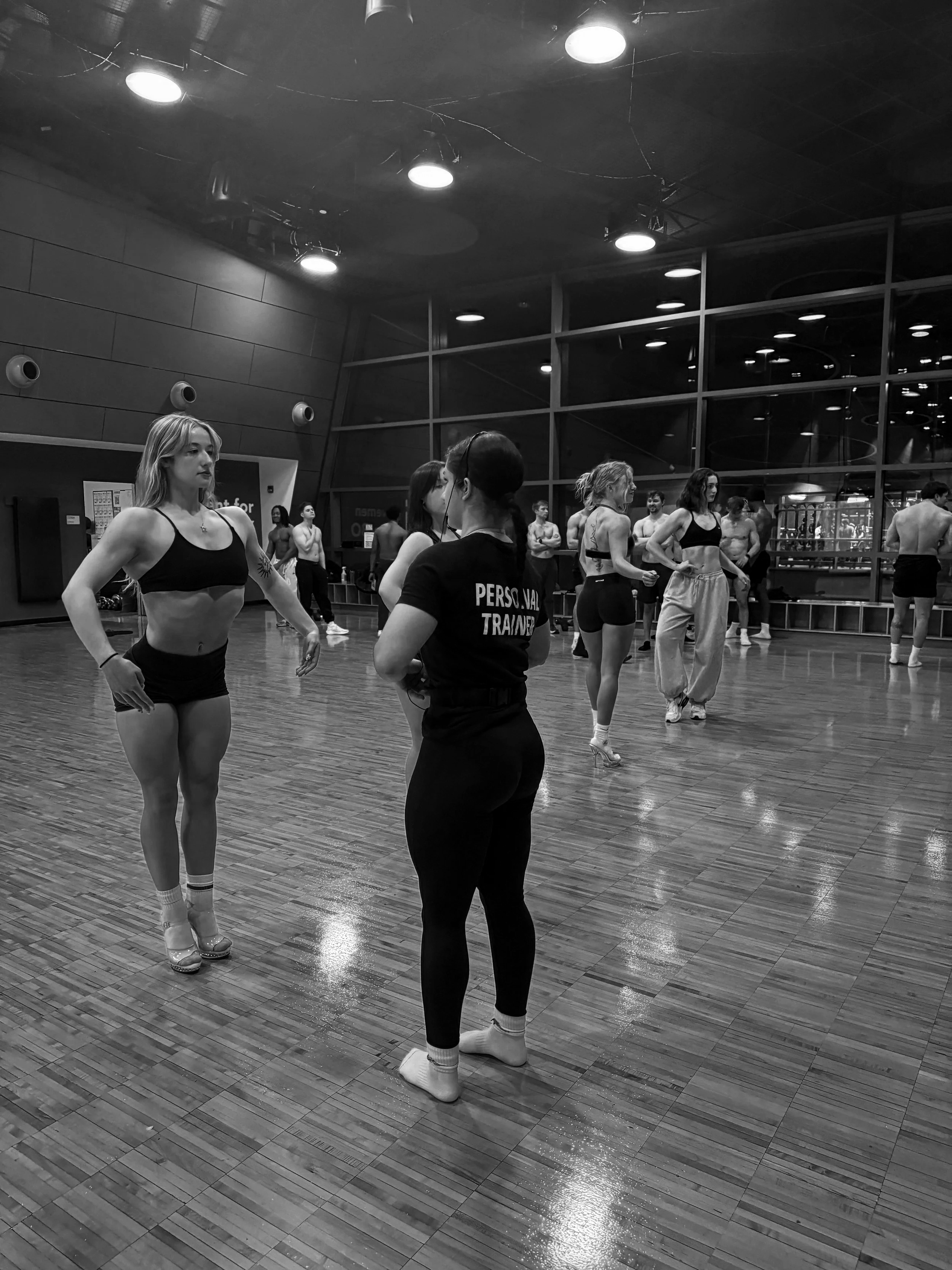 A group of people in workout attire, including women with dance shoes, in a gym or dance studio with wooden floors, large windows, and ceiling lights, preparing for a dance or fitness class.