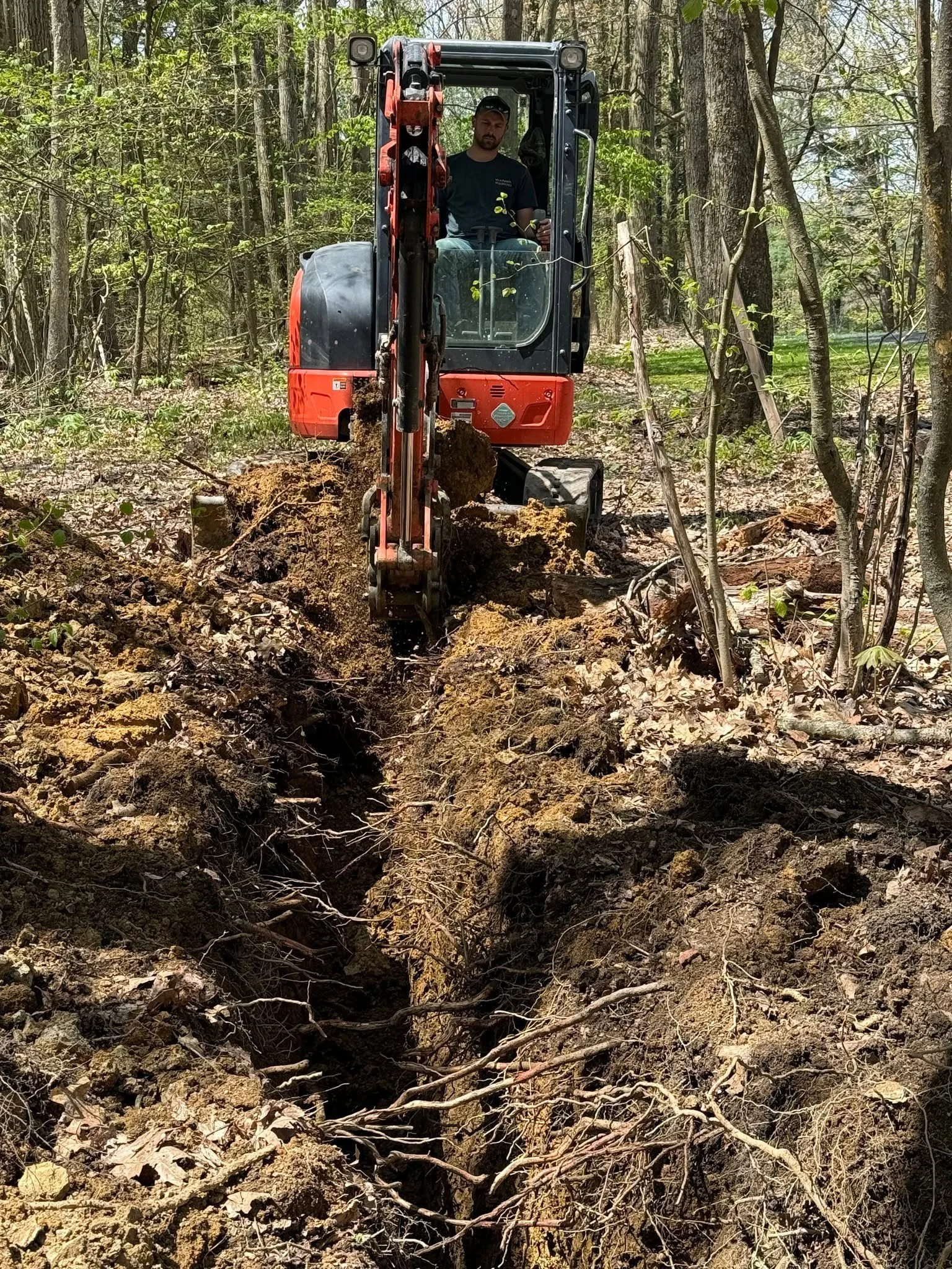 A man operating a small excavator digging a trench in a wooded area with trees and dirt.