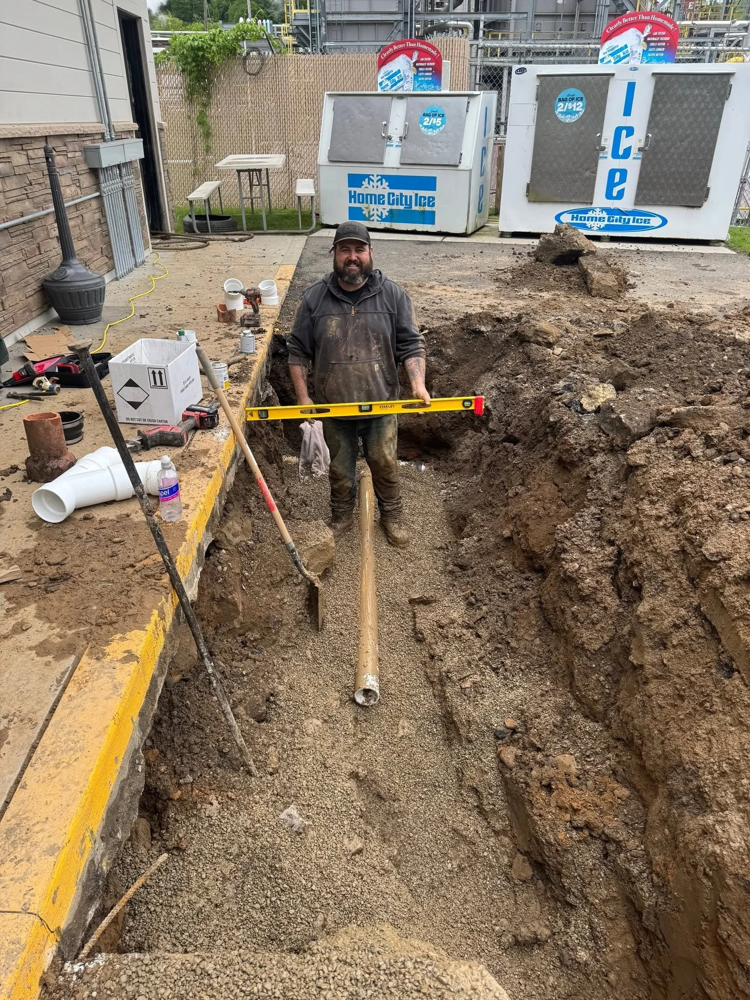 A construction worker standing inside an excavated trench with construction tools and materials around him, holding a yellow level, with two large ice storage units labeled 'Home City Ice' in the background.