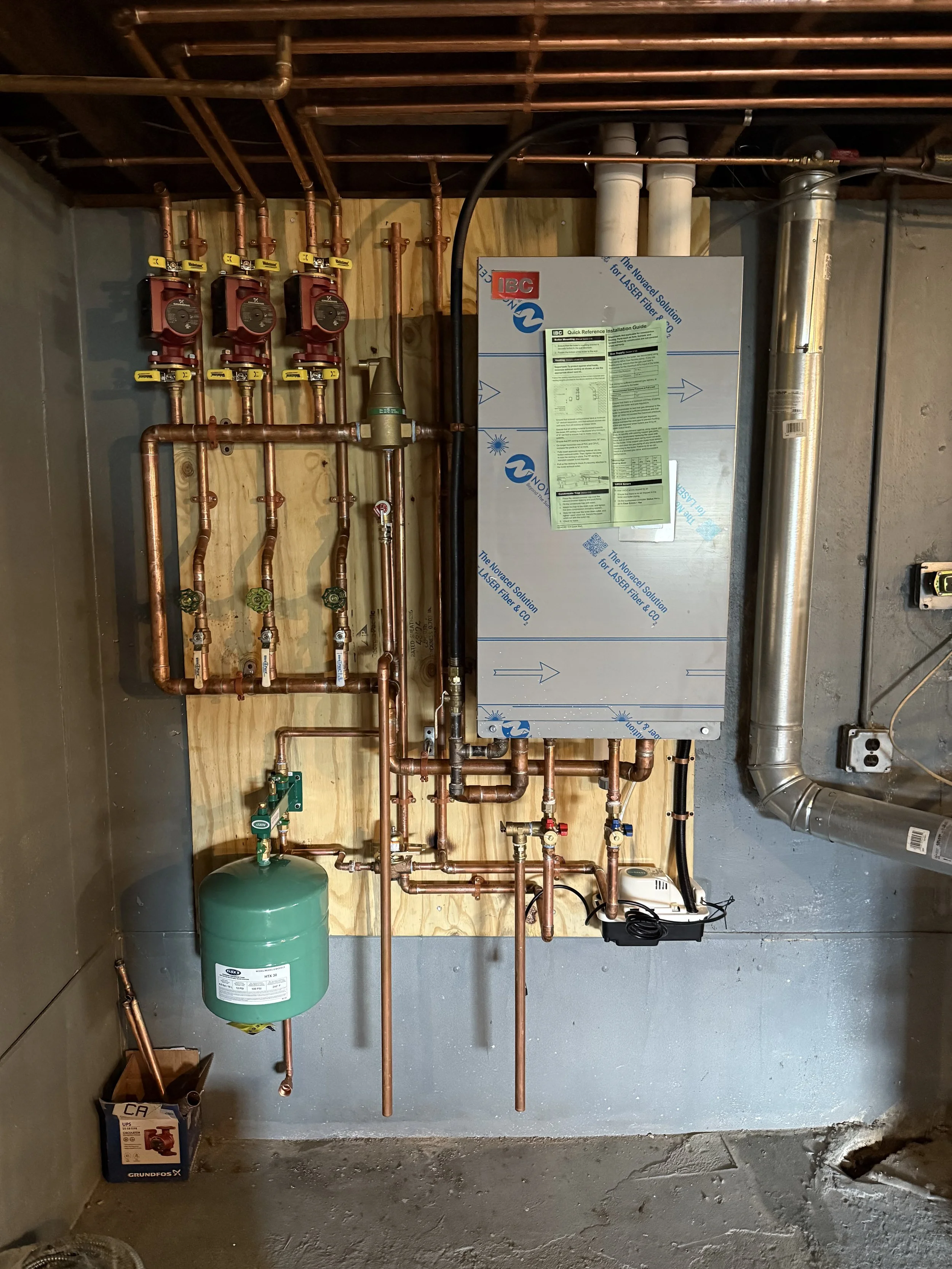 A utility room with copper pipes, valves, a water pressure gauge, a green expansion tank, and a gray control panel for a heating or plumbing system, installed against a wooden wall.