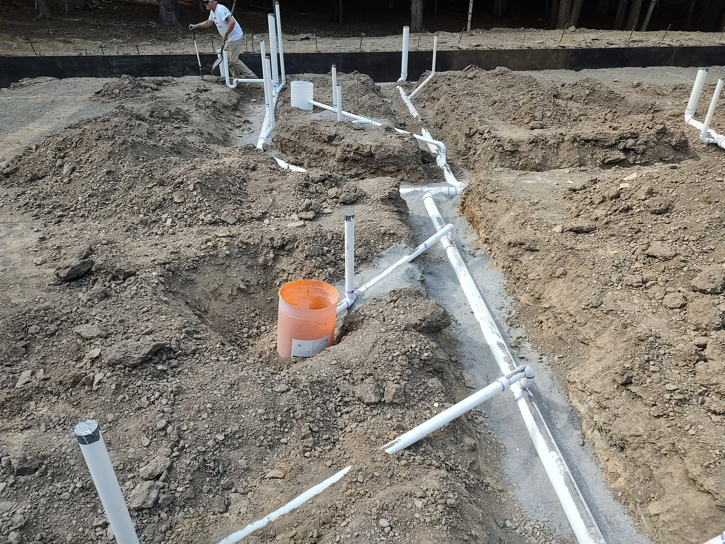 Construction site with exposed dirt and white PVC pipes being installed for plumbing or drainage system, with a worker in the background.