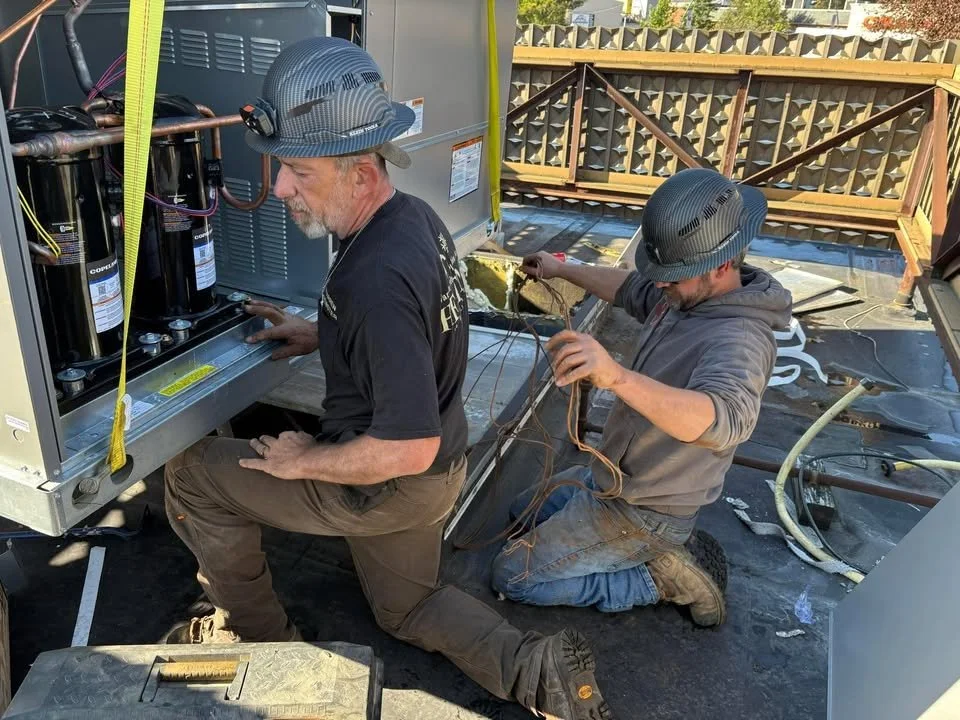 Two technicians working on HVAC or refrigeration units on a rooftop, wearing safety helmets, with tools and wires, with a wooden railing and trees in the background.