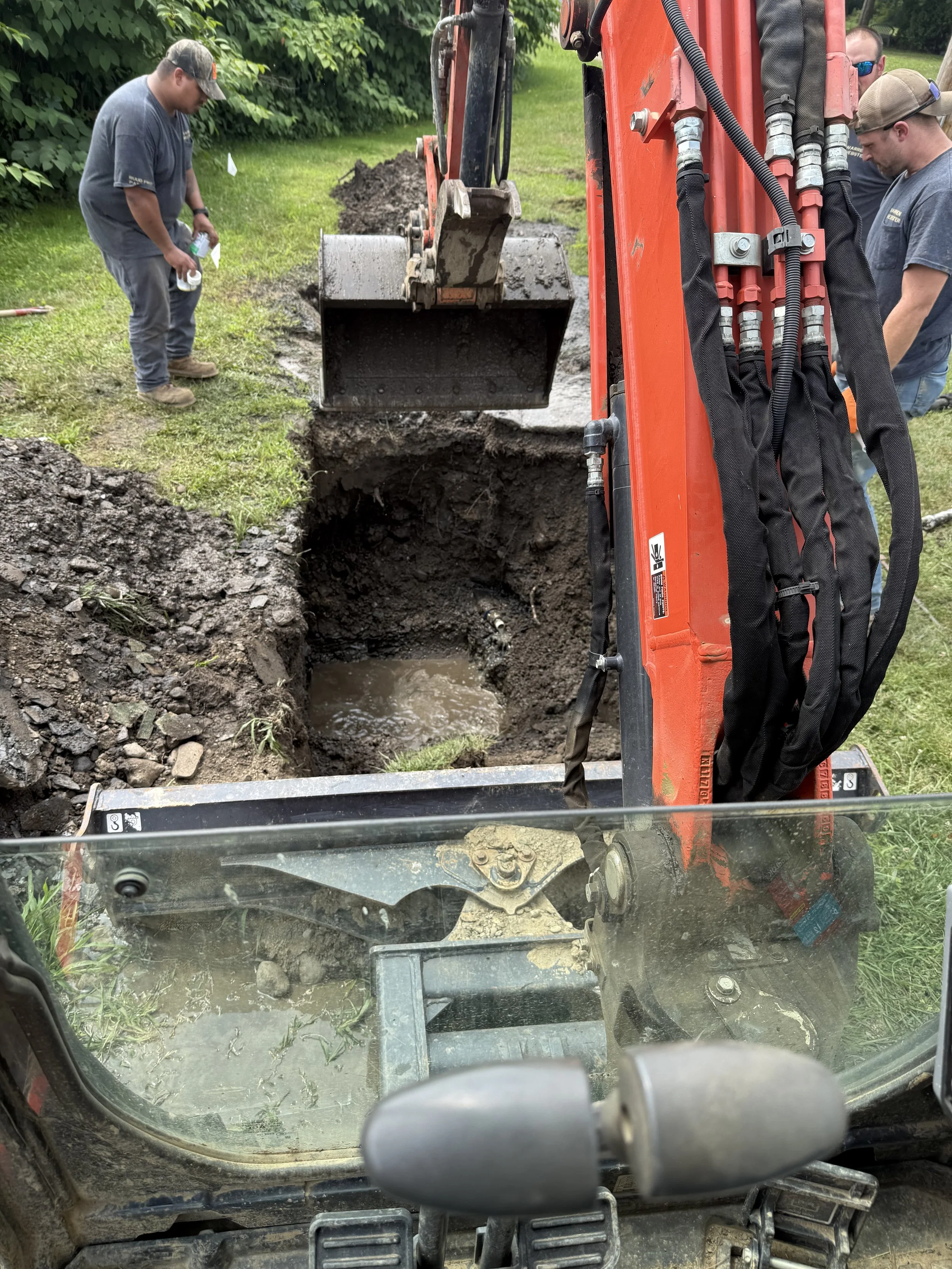 View from inside a construction vehicle showing an excavator digging a trench with three workers standing nearby, working outdoors on a grassy area with trees in the background.