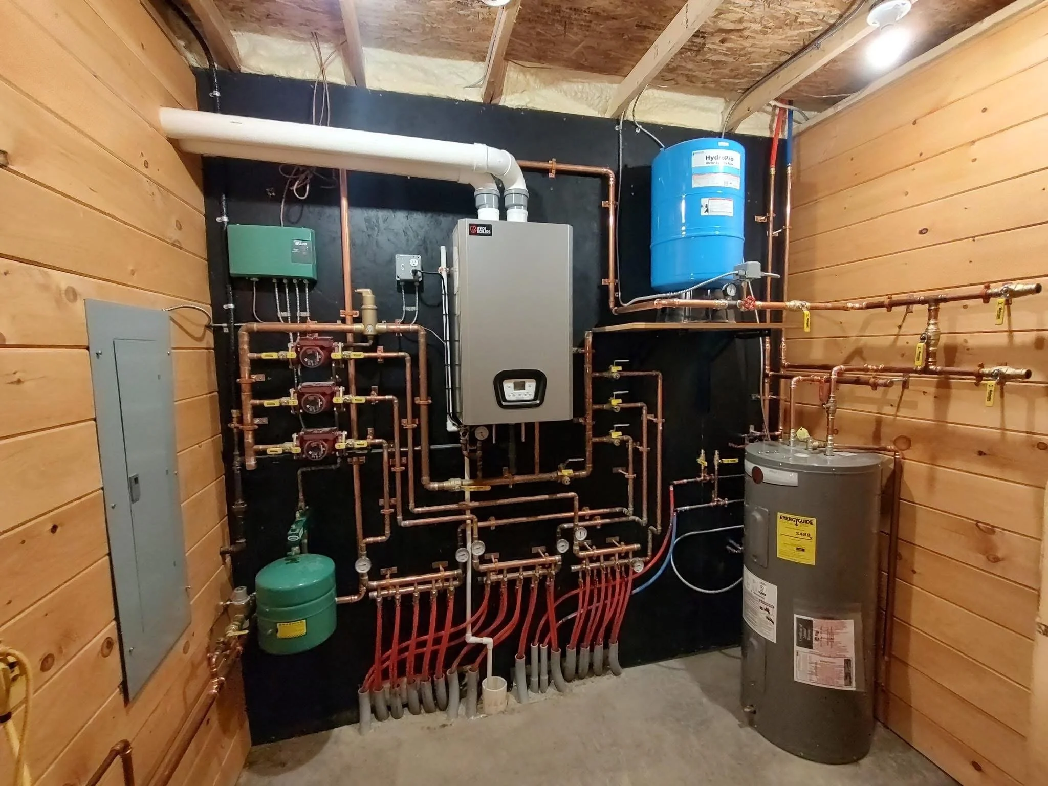 A utility or mechanical room with copper piping, a water heater, and a water pressure tank, built with wood-paneled walls and a black background wall.