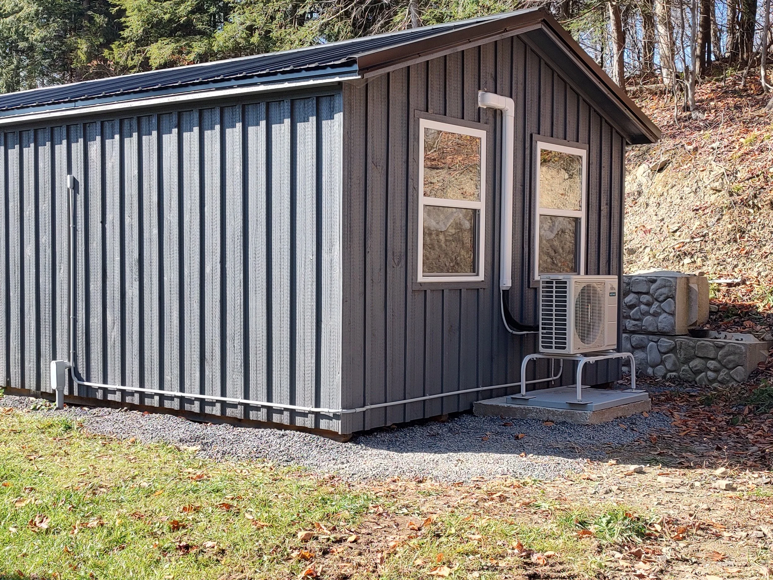 A small, vertical metal building with a fieldstone foundation, vertical wood siding, and two small windows, situated outdoors with trees and fallen leaves in the background.