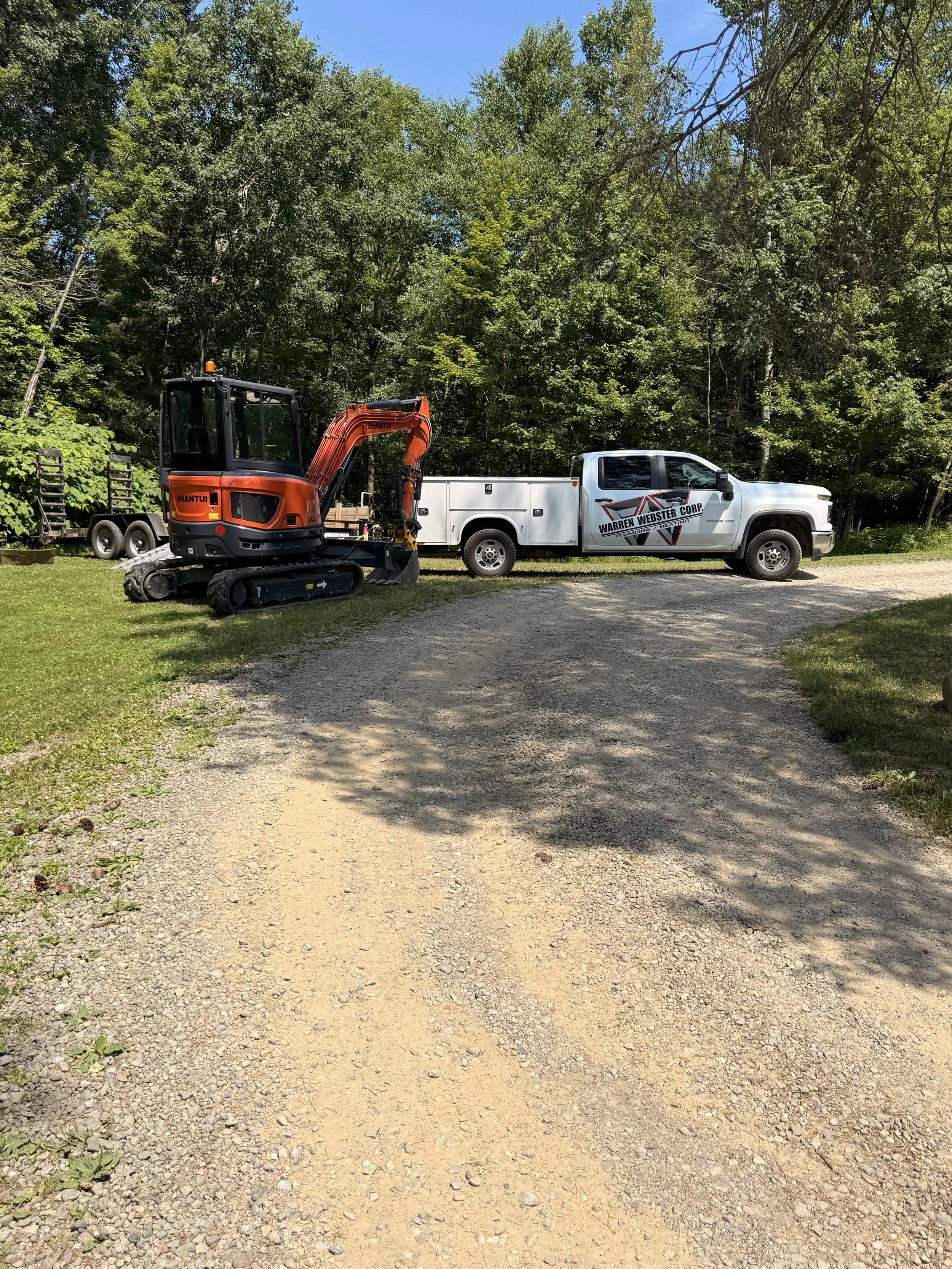 A white work truck with the logo 'Warren Webster Corp.' parked on a gravel driveway beside a small orange excavator with the brand name 'Shantui'. There are trees and a blue sky in the background.