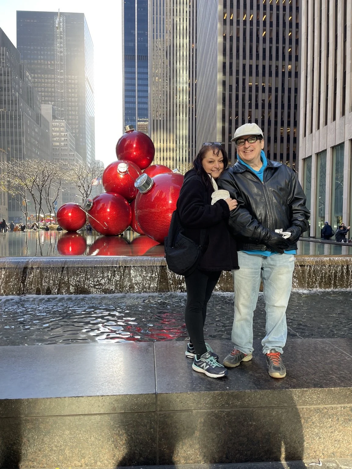 Two people standing together at Rockefeller Center in New York City, with large red Christmas ornaments and a water fountain behind them, surrounded by tall skyscrapers.