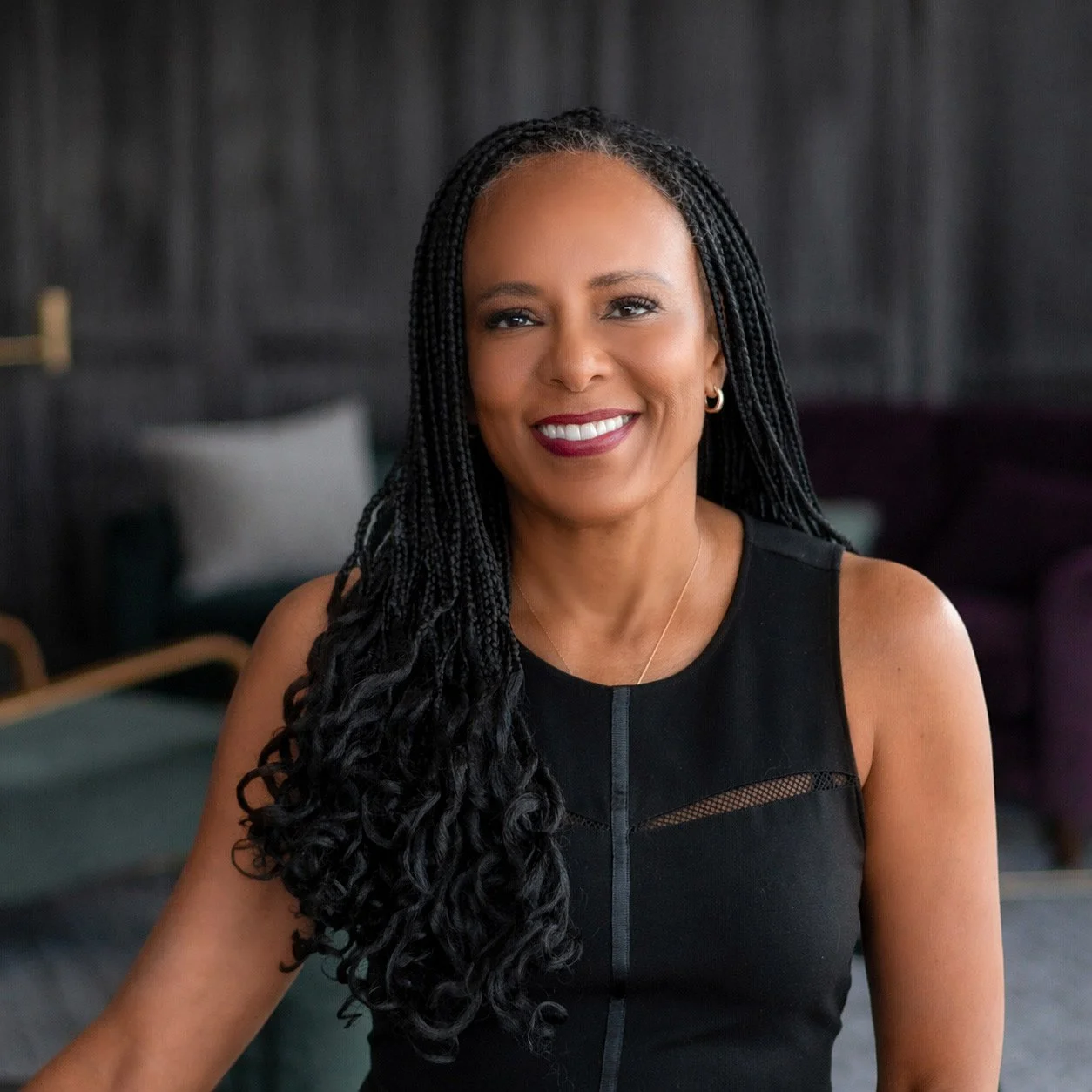A smiling woman with long black braided hair, wearing a sleeveless black dress, sitting in a modern, stylish room with dark walls and comfortable furniture.