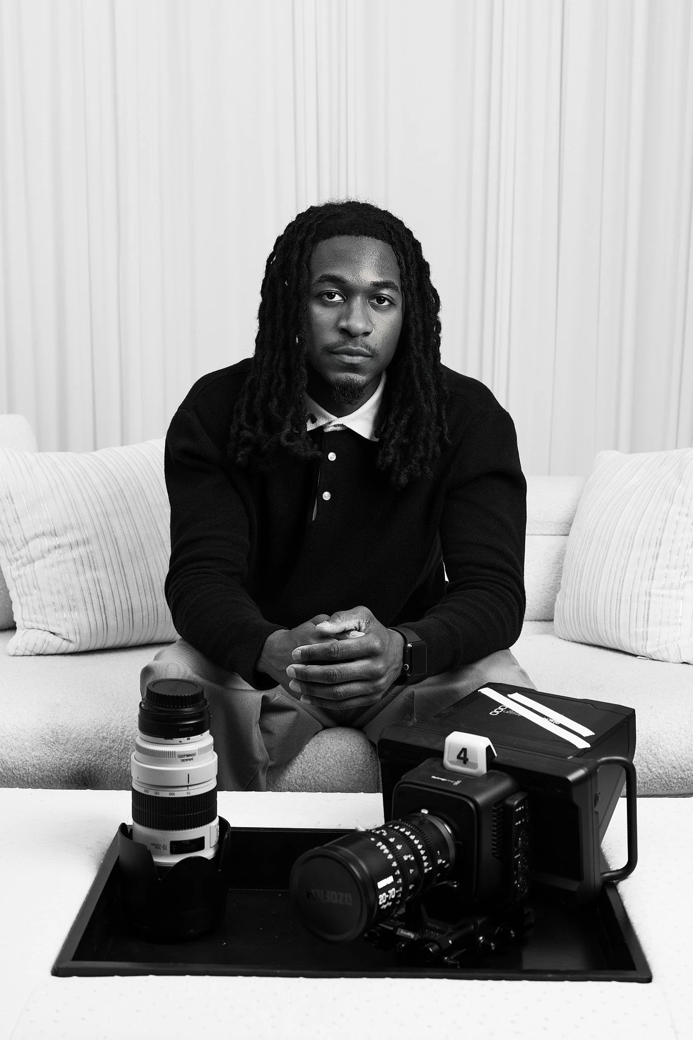 A man with dreadlocks sitting on a sofa with cameras, lenses, and photography accessories in front of him in a room with white curtains.