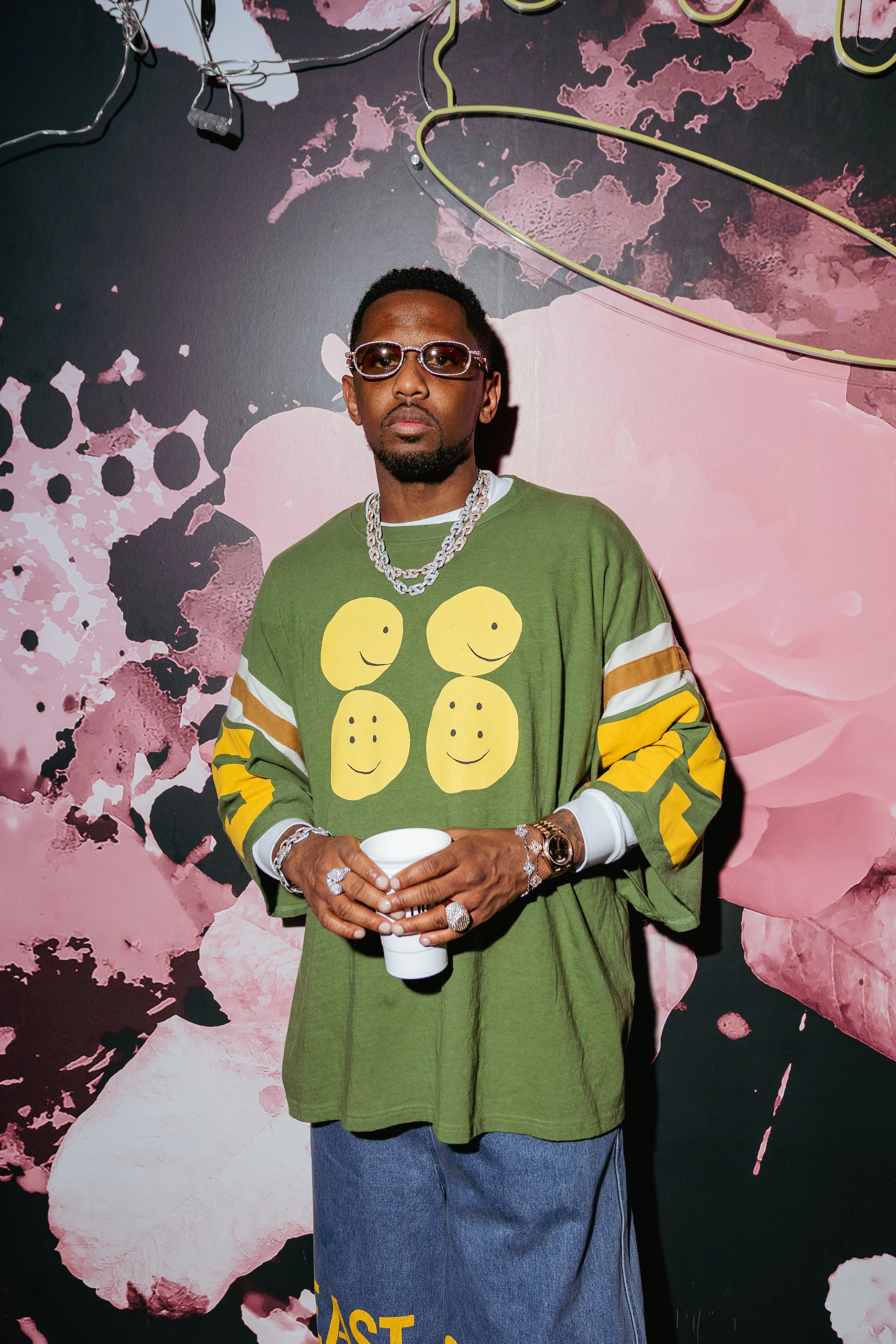 A man wearing glasses, a large silver chain, and a green sweatshirt with yellow smiley faces stands in front of a pink and black abstract background, holding a white cup.