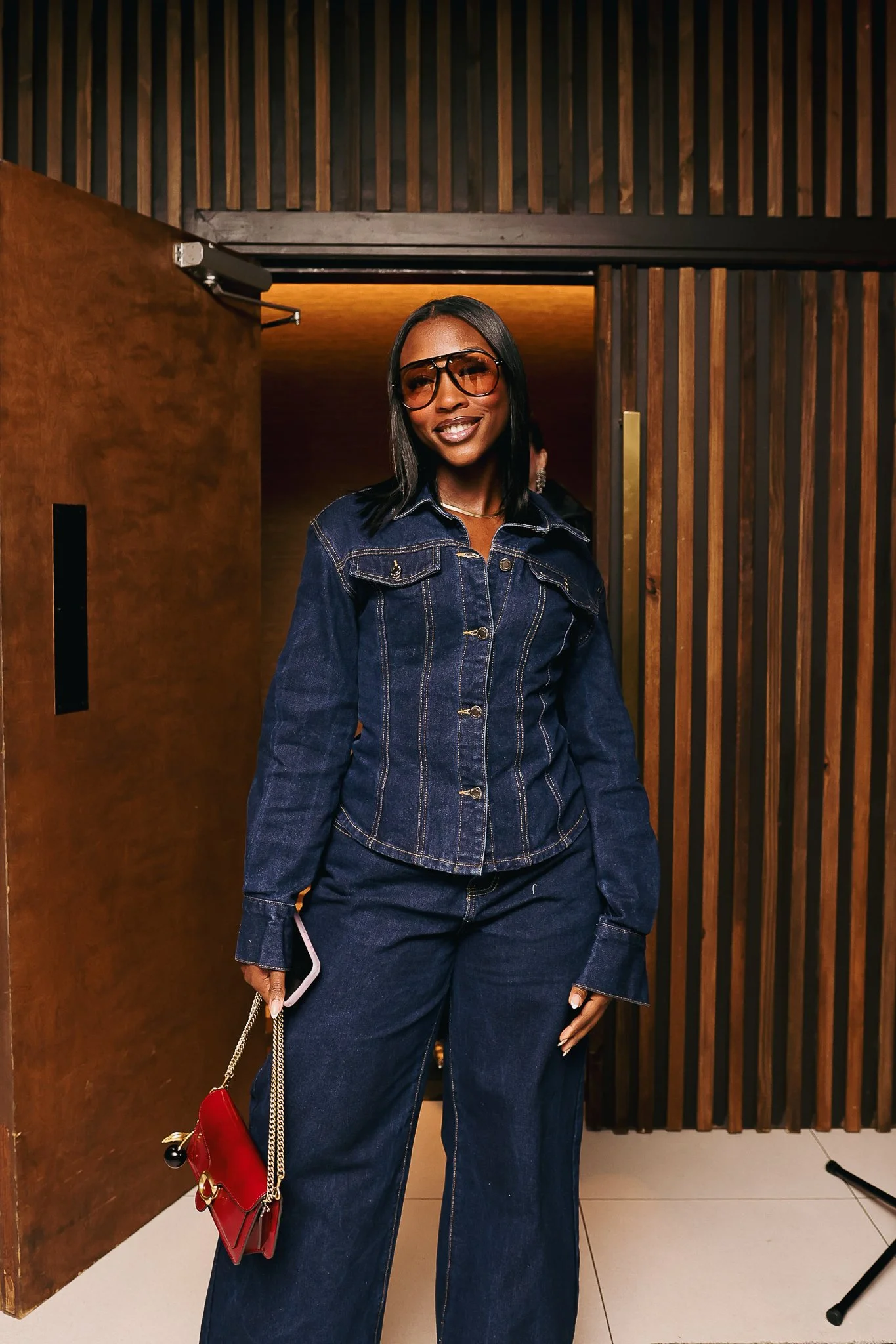 A woman standing in an indoor space with wooden paneling on the wall, wearing large sunglasses, a denim jacket, matching denim pants, and holding a red handbag with a gold chain strap, smiling at the camera.