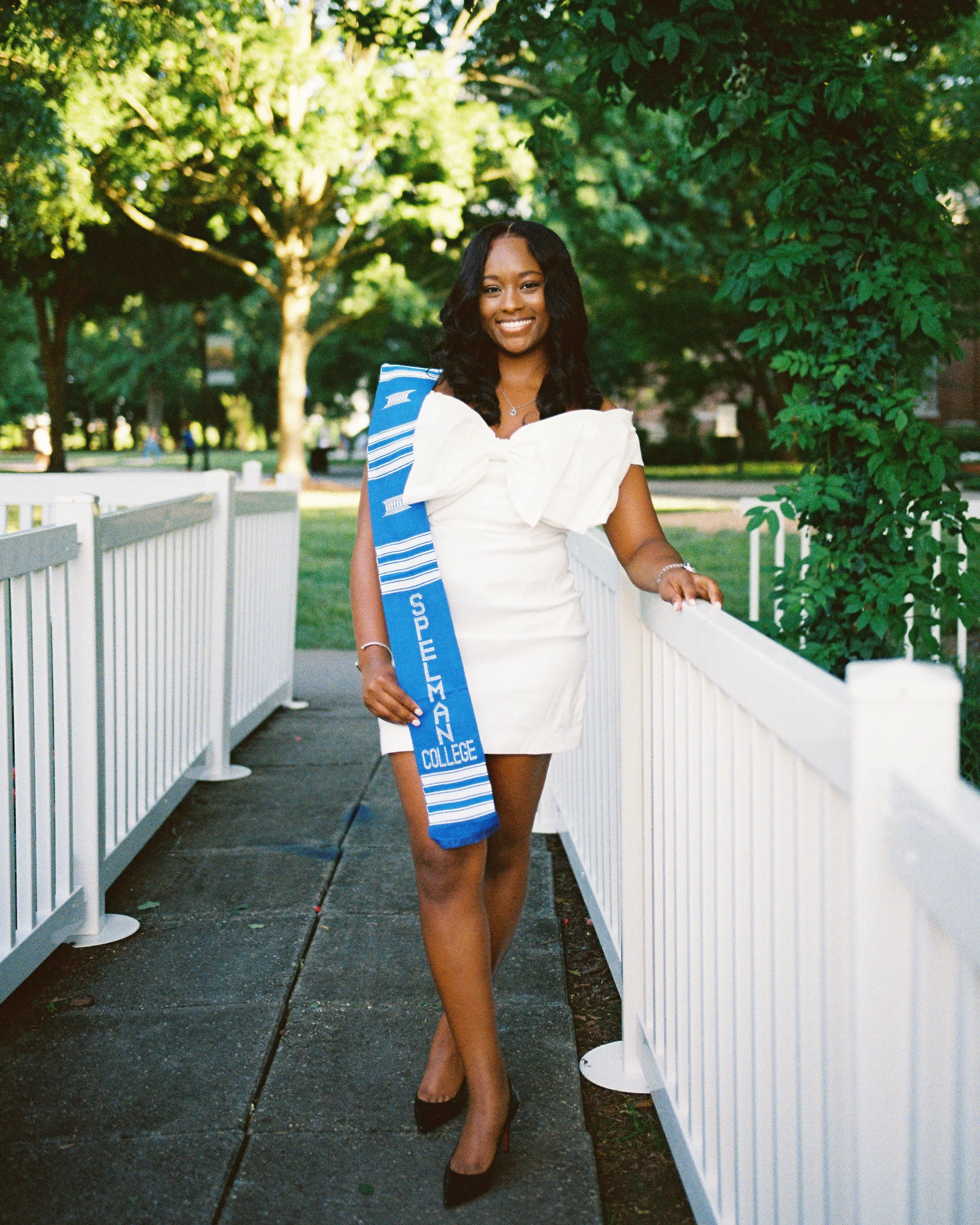 A young woman in a white dress and high heels standing outdoors on a paved walkway with a white picket fence on one side and green trees in the background. She is smiling and wearing a blue and white sash.