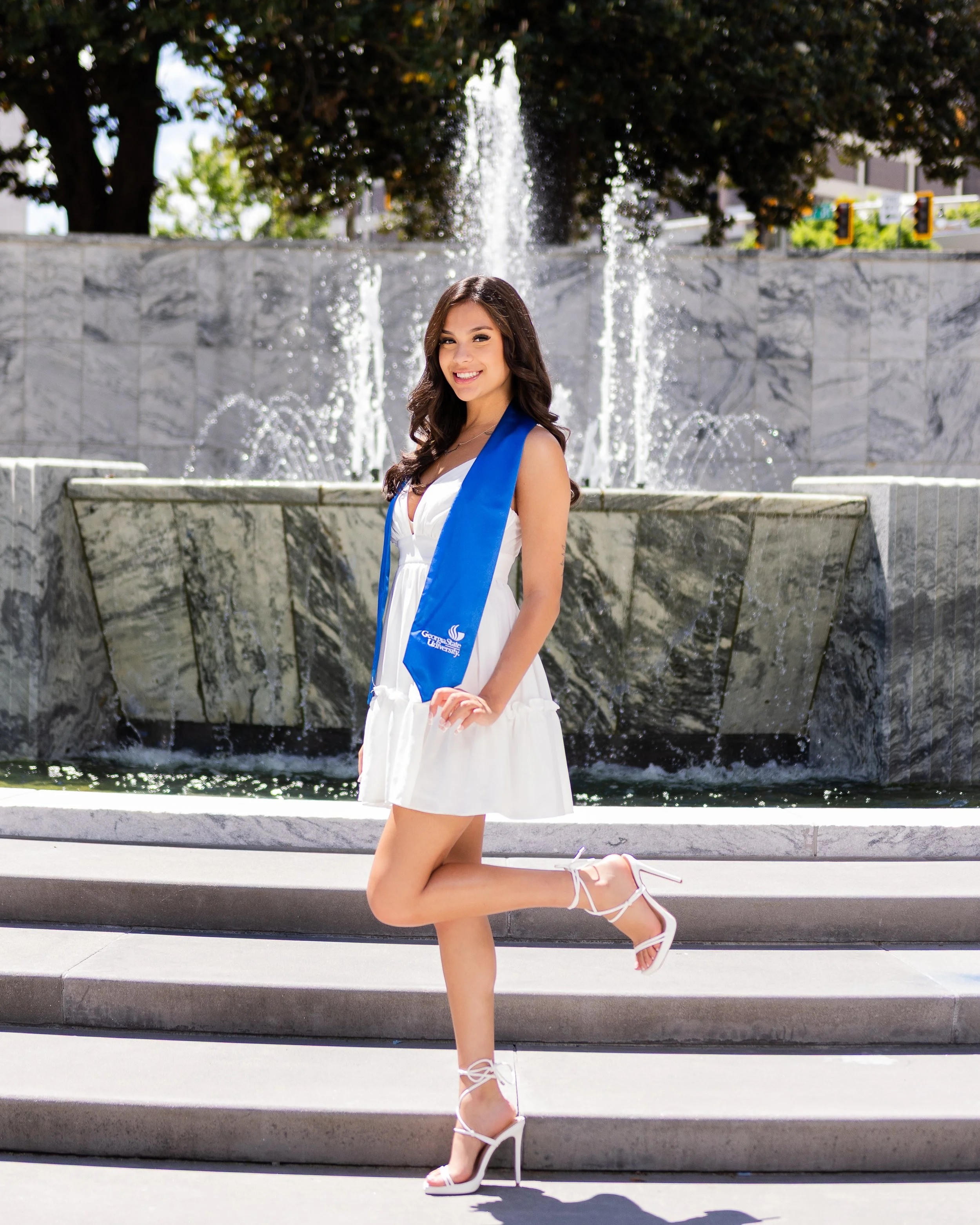 Young woman in white dress with blue sash, kneeling on stairs in front of a water fountain, smiling at the camera.