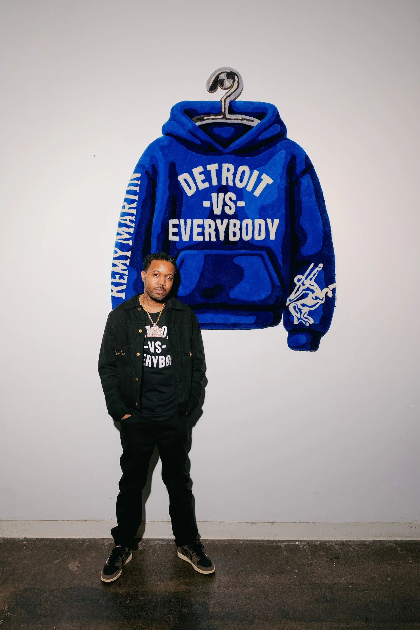 A man standing in front of a wall with a painting of a blue hoodie that reads 'Detroit vs Everybody' with a figure's face and a hand holding a gun on the sleeve, wearing black clothing and sneakers.