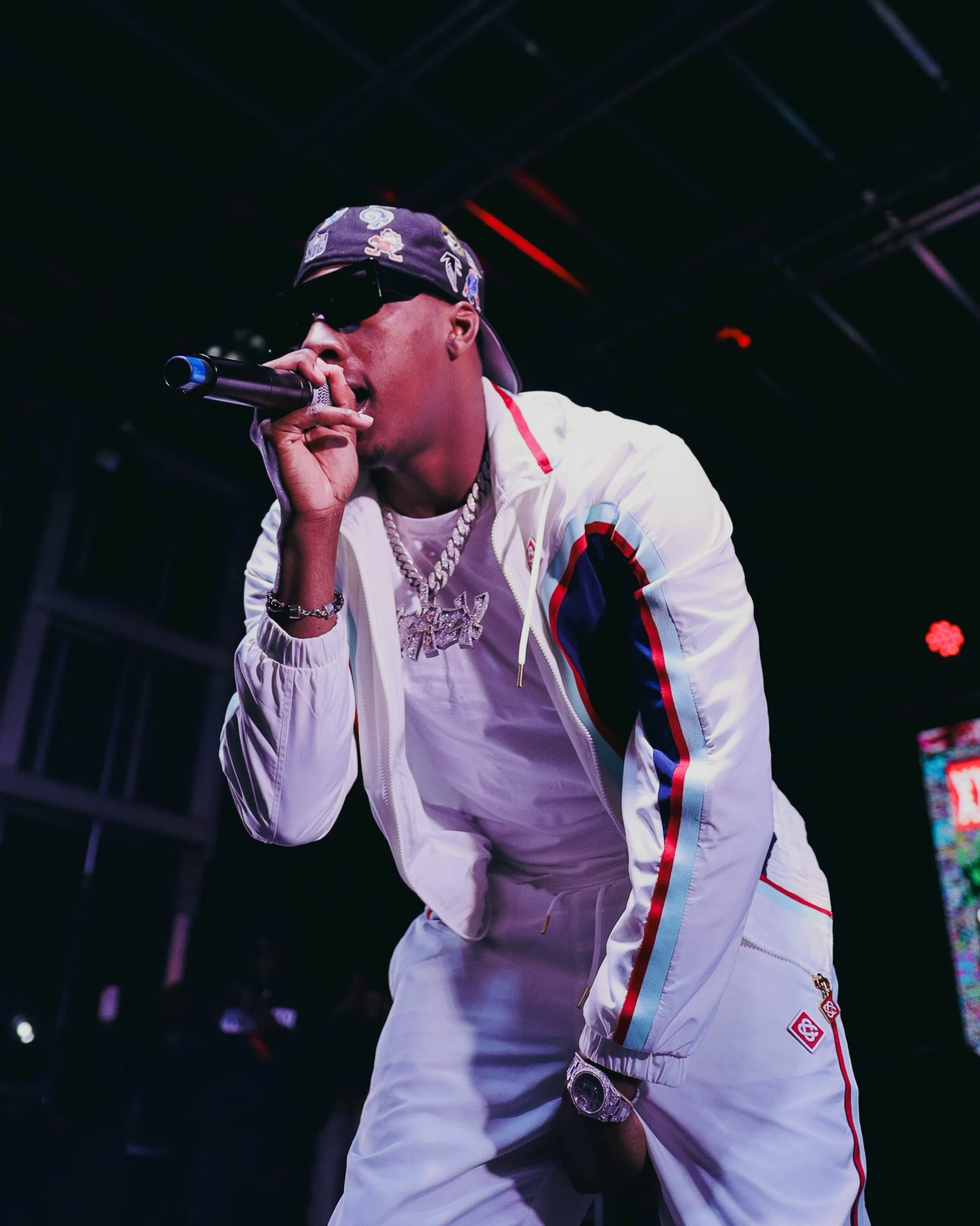 A rapper wearing dark sunglasses, a bandana with patches, a white track jacket with red and blue stripes, and jewelry, performing on a stage with a microphone in hand.