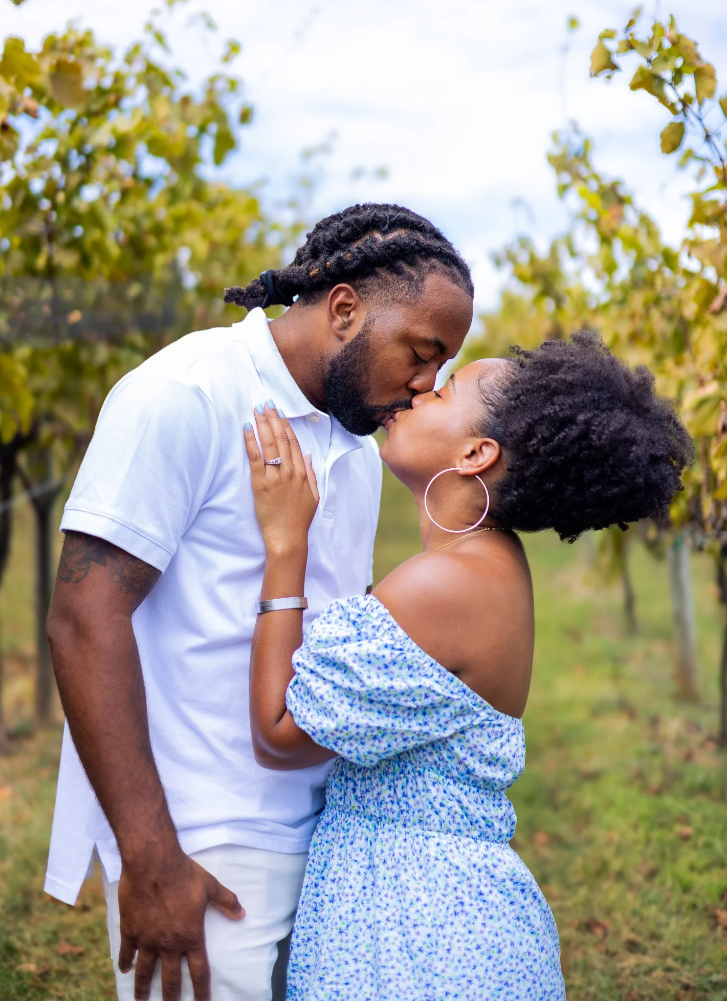 A couple sharing a kiss outdoors, standing close with eyes closed. The man has dreadlocks and a beard, wearing a white polo shirt. The woman has natural curly hair, wears large hoop earrings, and a blue patterned dress. The background shows green tre
