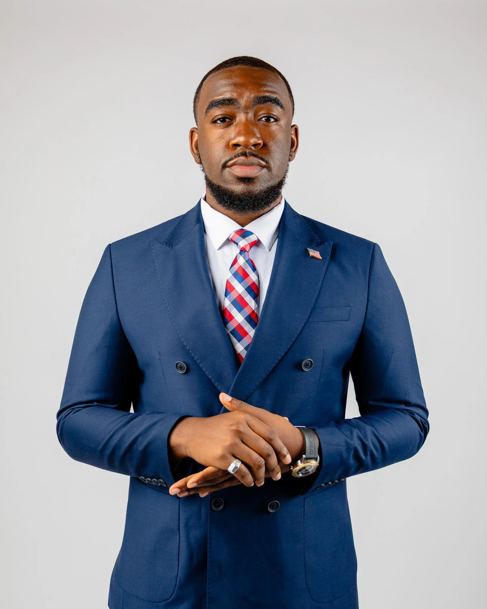 A man in a blue suit with a red, white, and blue plaid tie and a small American flag lapel pin, standing against a plain gray background.