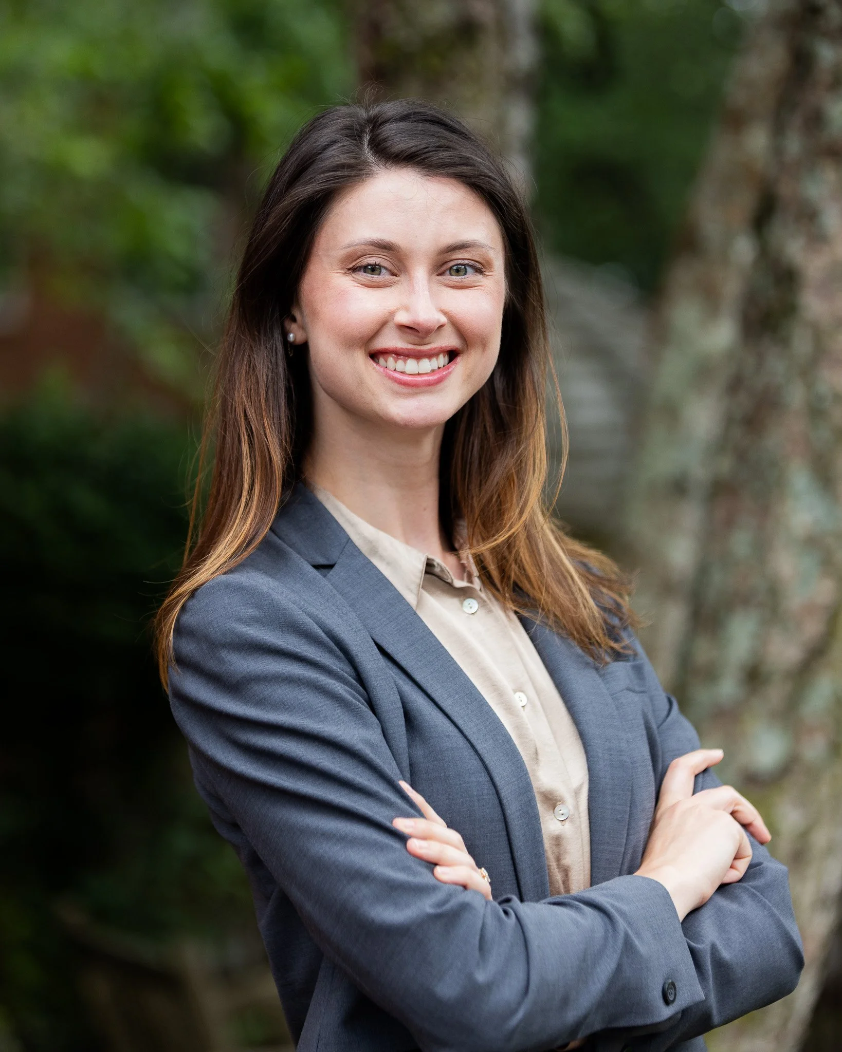 A woman with long brown hair, wearing a gray blazer and a beige blouse, standing outdoors with blurred greenery in the background, smiling and crossing her arms.