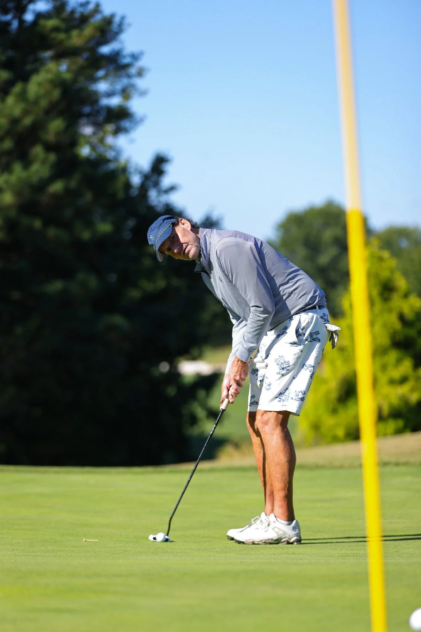 A man on a golf course preparing to putt a golf ball near the hole, with the flagstick visible in the background, trees along the course, and a clear blue sky.