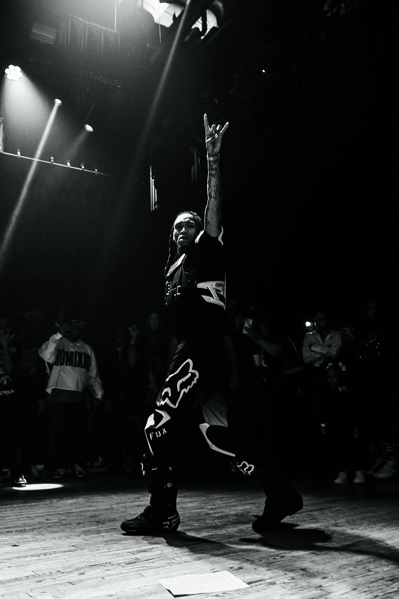 A person with long hair dancing on stage with one arm raised, surrounded by an audience, in a dark setting with stage lighting.