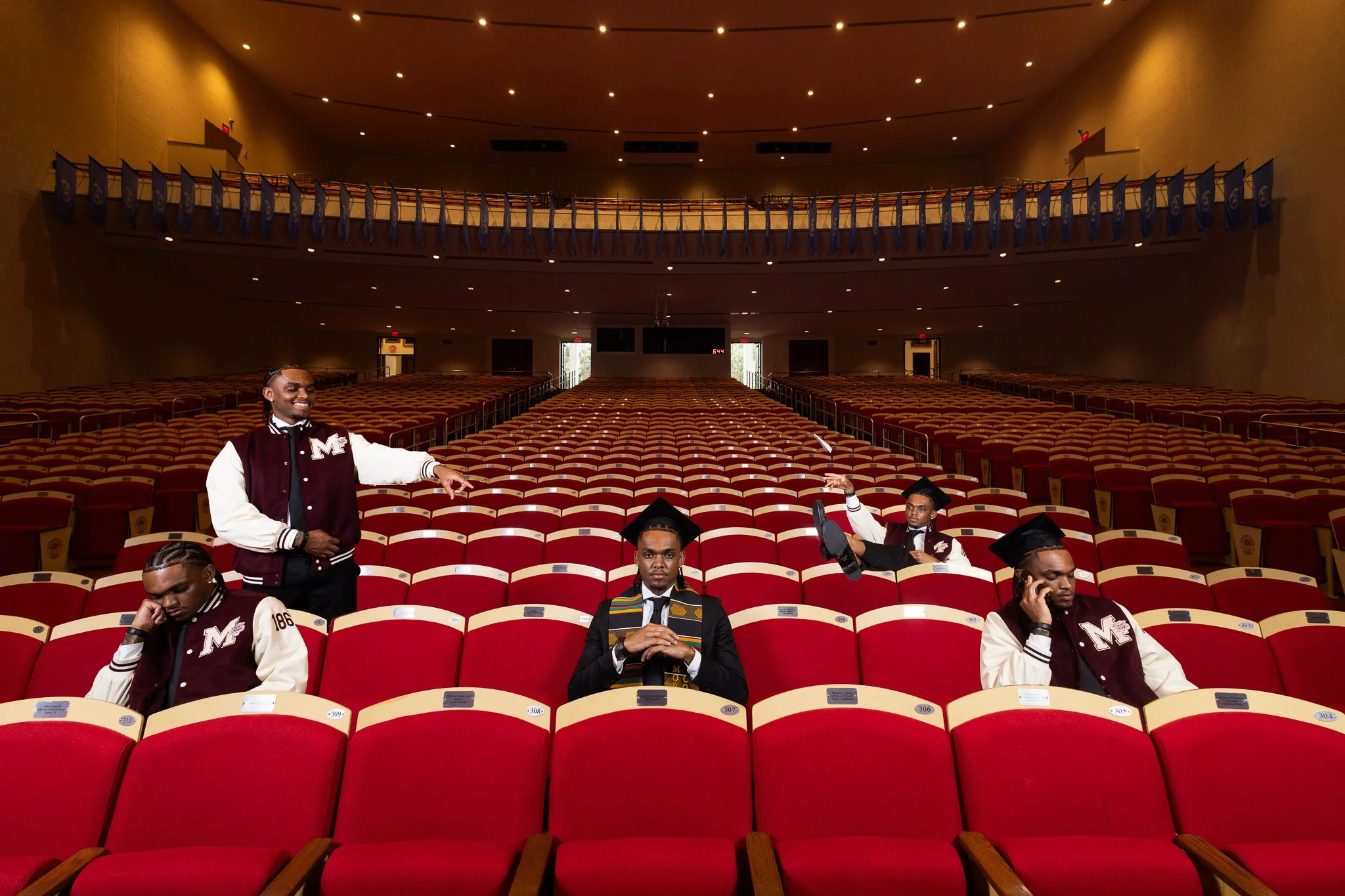 A large auditorium with rows of red seats and some students dressed in graduation gowns and caps, some sitting and some standing. The auditorium is mostly empty.