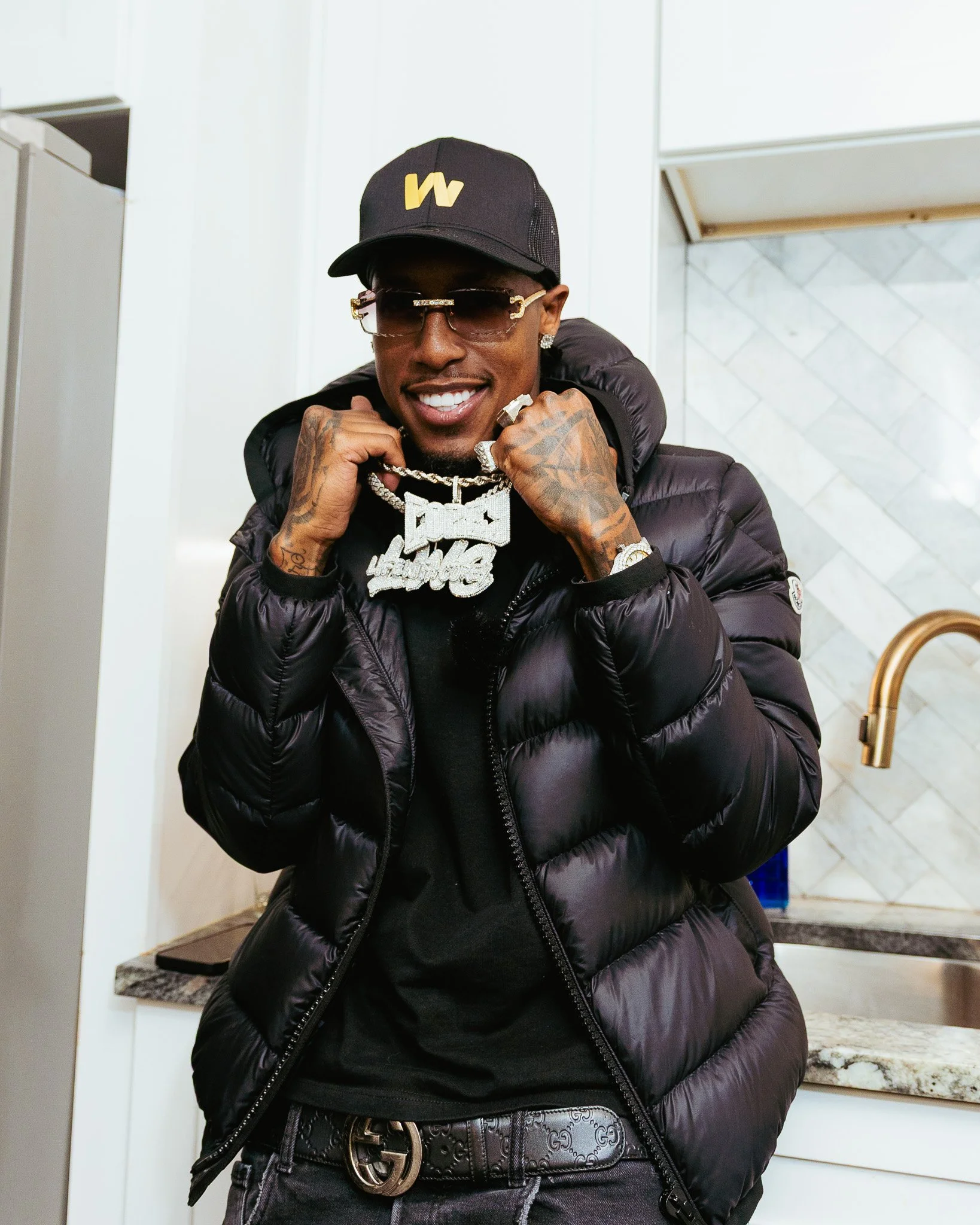 A smiling man wearing sunglasses, a black cap with a yellow logo, a black puffer jacket, and necklaces, standing in a modern kitchen with white cabinets and a marble countertop.