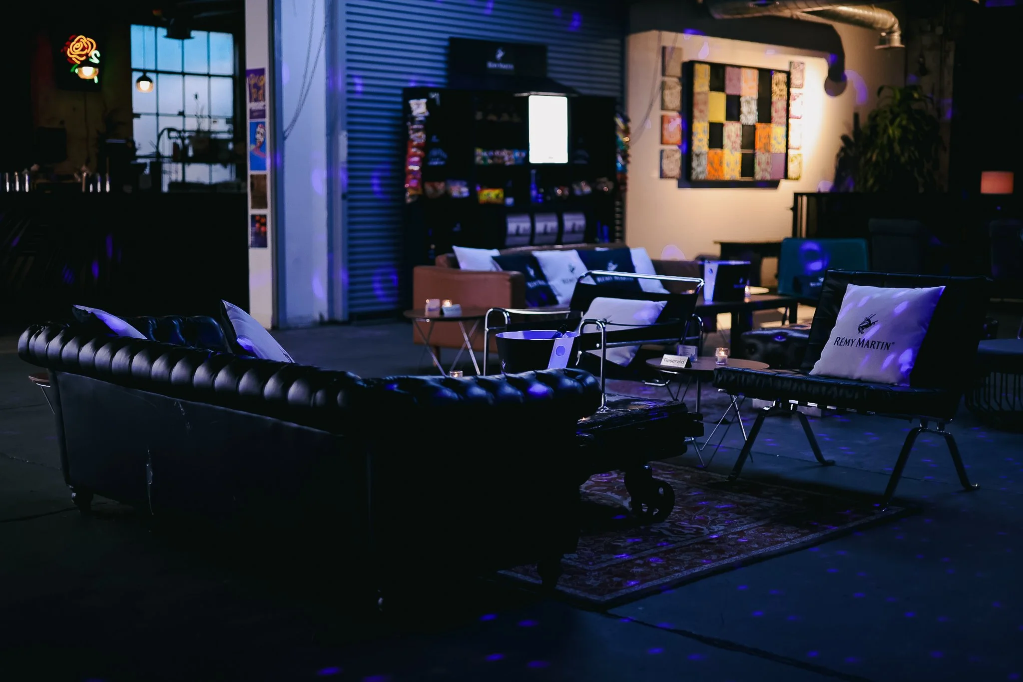 Dimly lit indoor lounge area with black leather couches, decorative pillows, small tables with candles, and wall art, illuminated by blue and purple lighting.