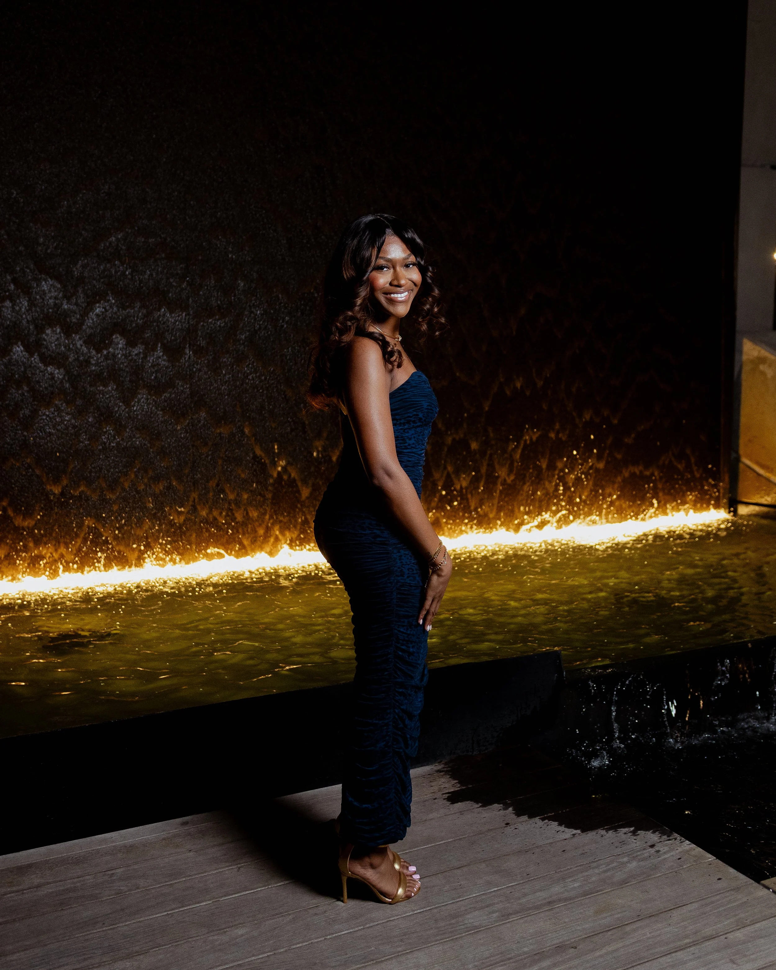 A woman in a strapless blue dress and gold high heels standing on a wooden floor in front of a dark textured wall with lighting at its base.