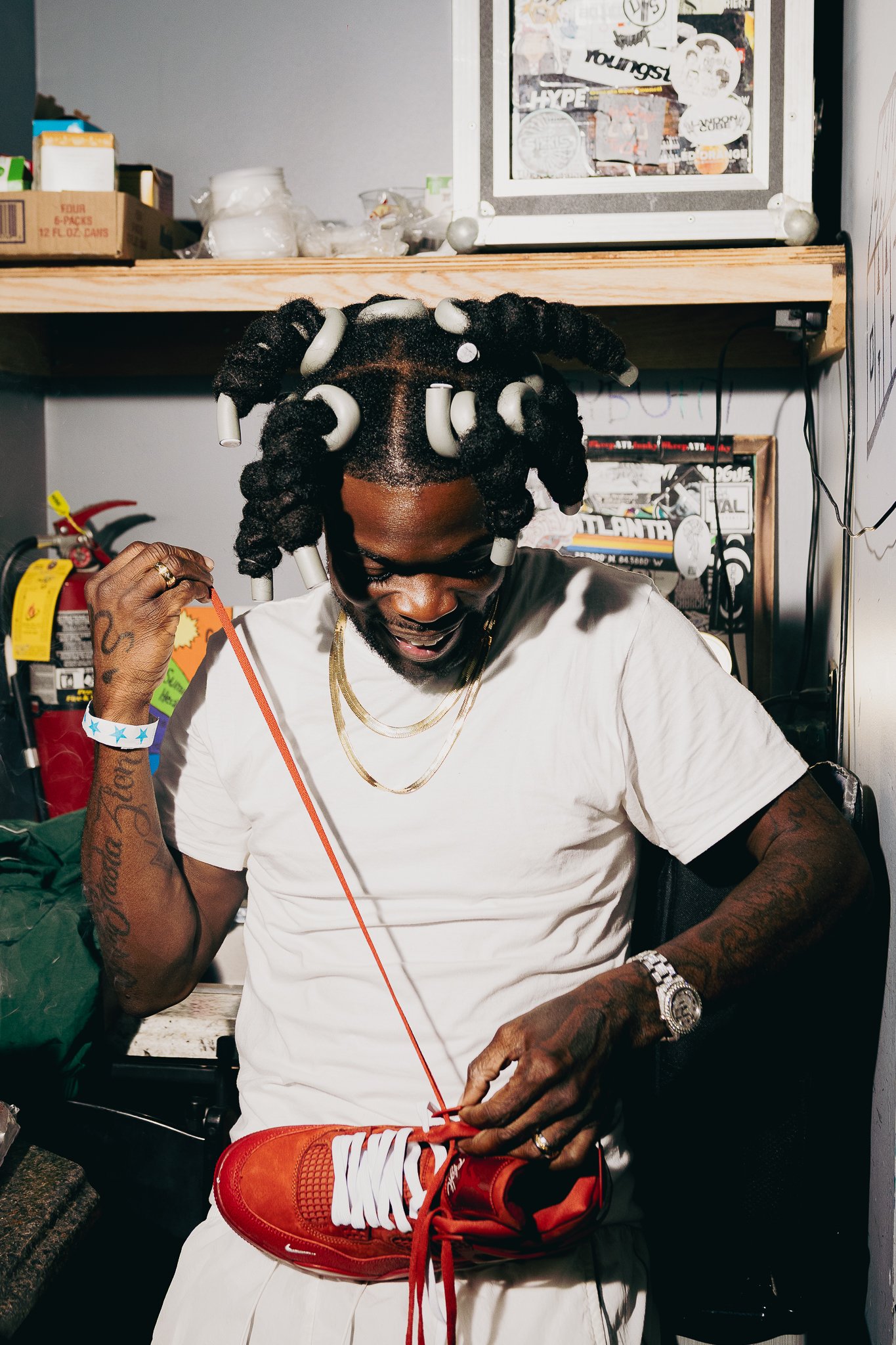 A man with textured hair wearing curlers, glasses, and jewelry is sitting and putting on red sneakers with white laces and detailing. He has tattoos on his arms, a watch, and a wristband. The background shows shelves with various items, including a b