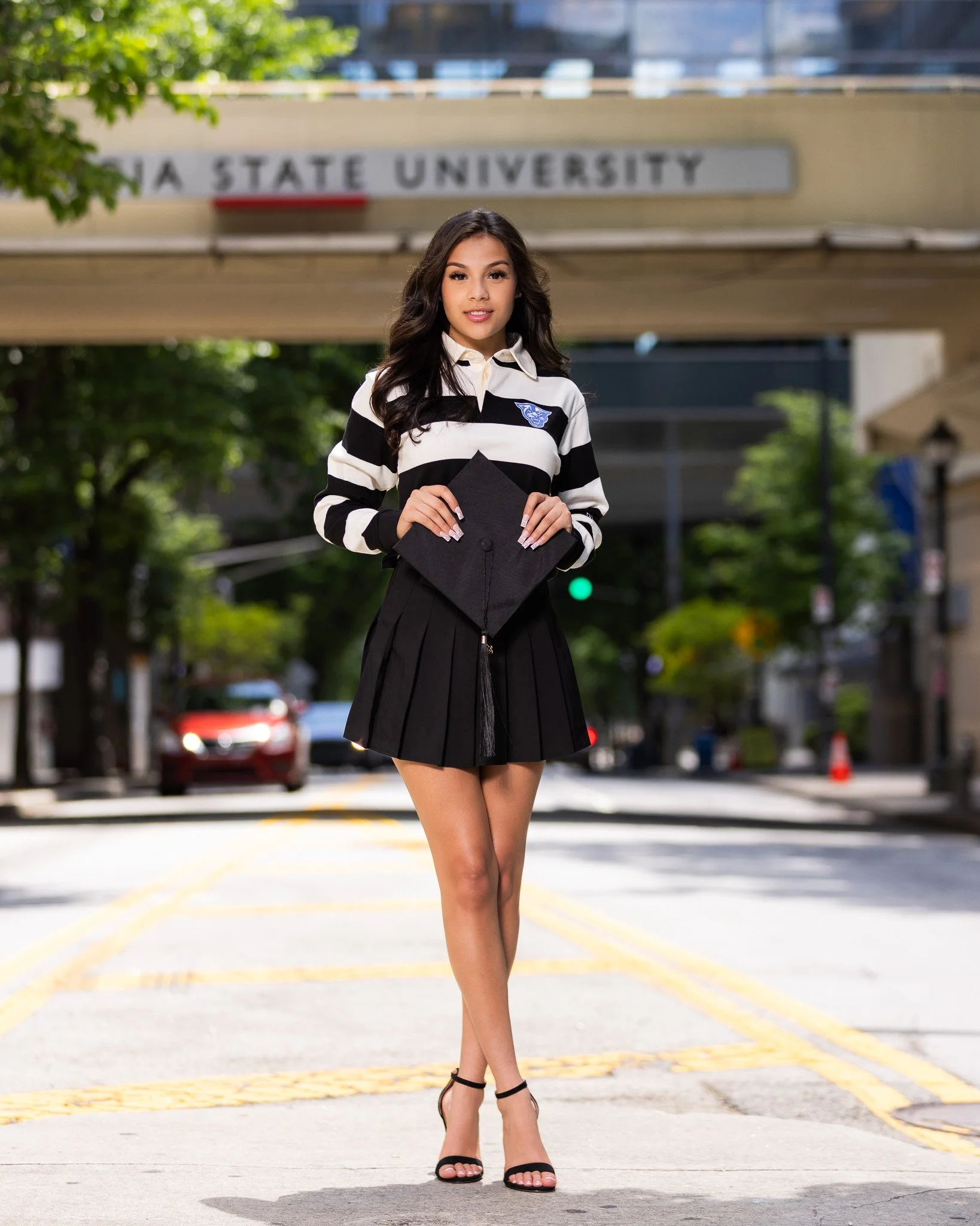 Young woman in a black and white striped shirt holding a graduation cap, standing on a city street with a university sign in the background.