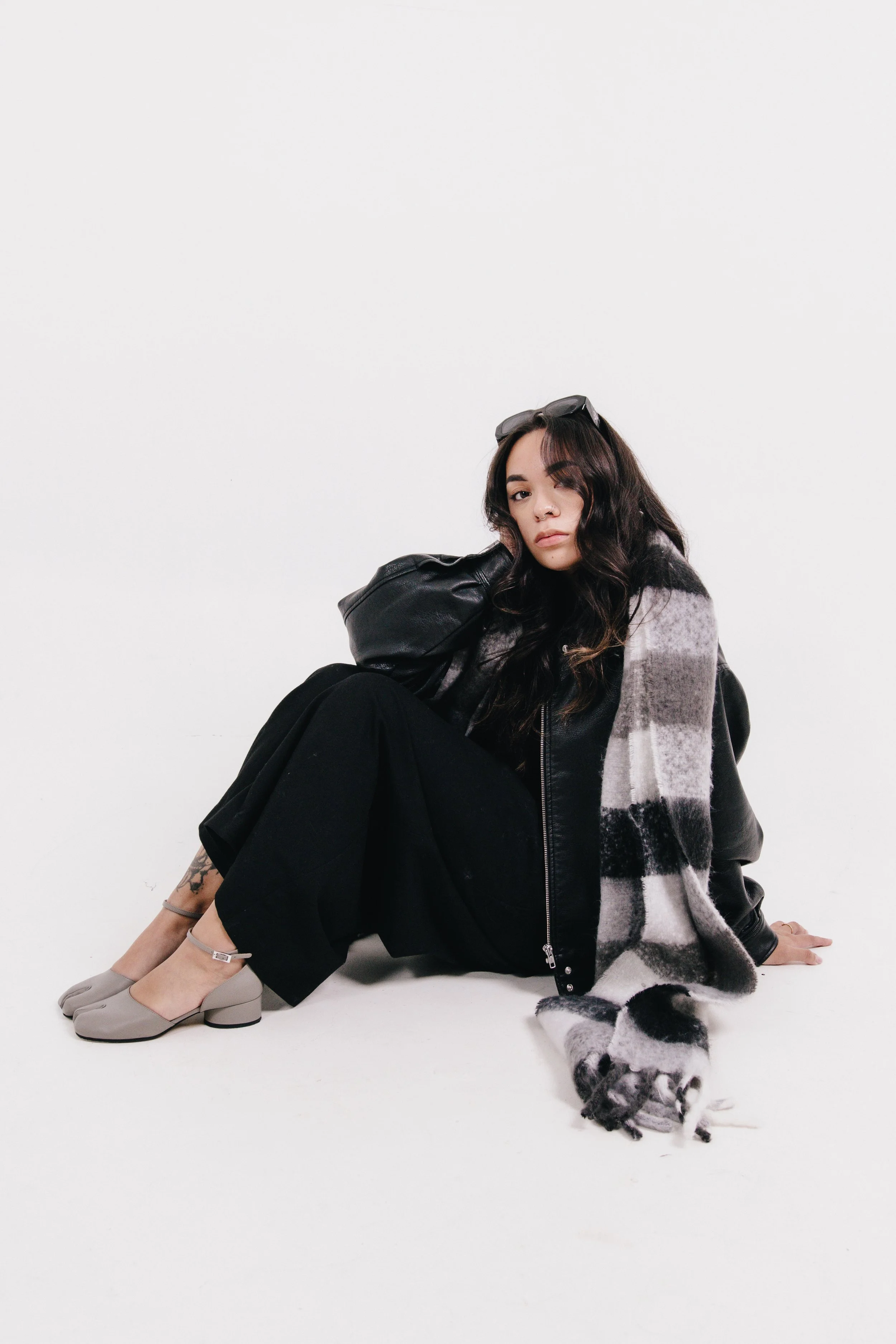 A woman with long dark wavy hair, sitting on the floor against a white background, wearing a black leather jacket, beige shoes, sunglasses on her head, and a plaid scarf, with a Dalmatian puppy lying next to her.