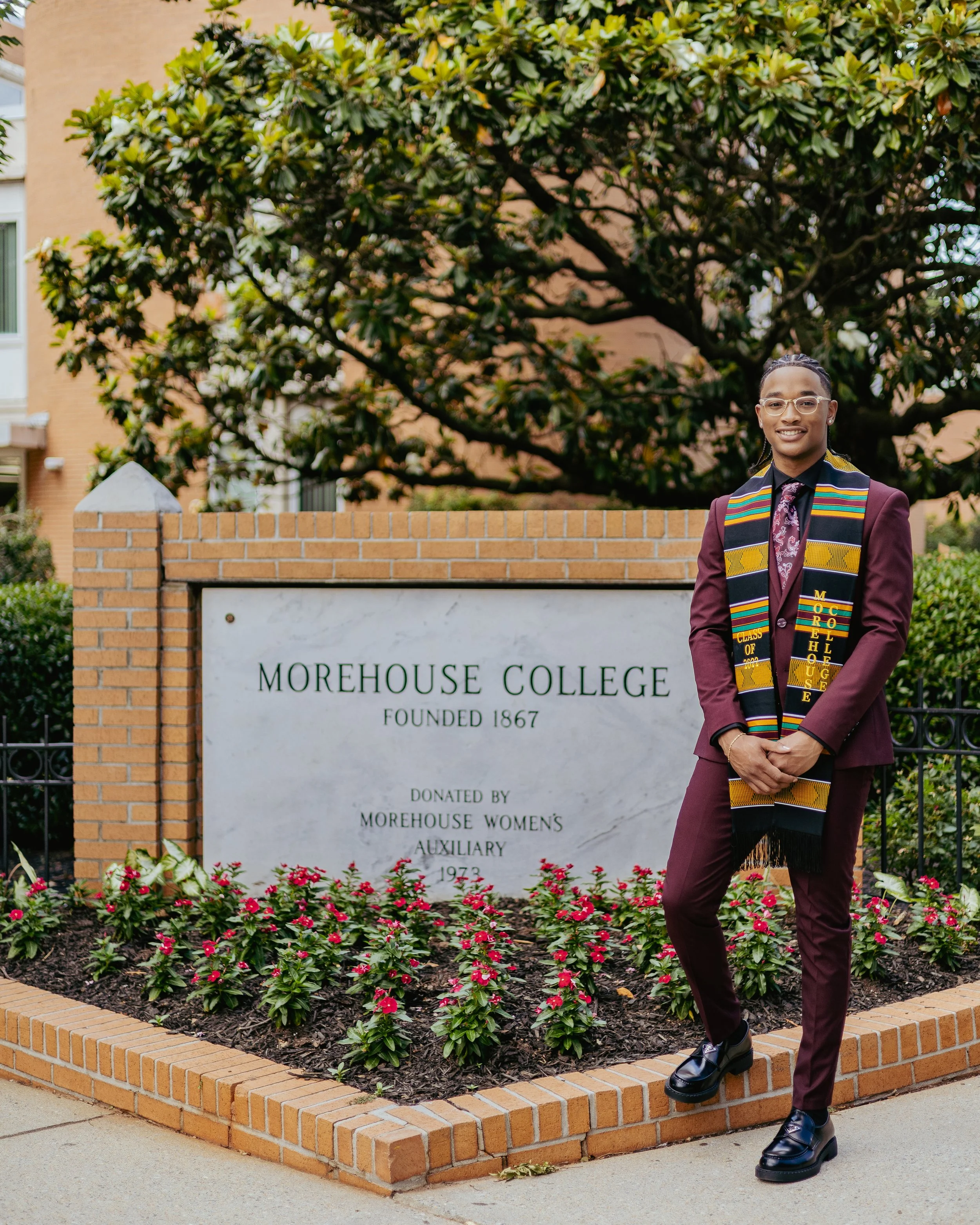 A young person wearing glasses, a dark purple suit, and a colorful graduation stole standing in front of a sign for Morehouse College with a brick border and flower bed.