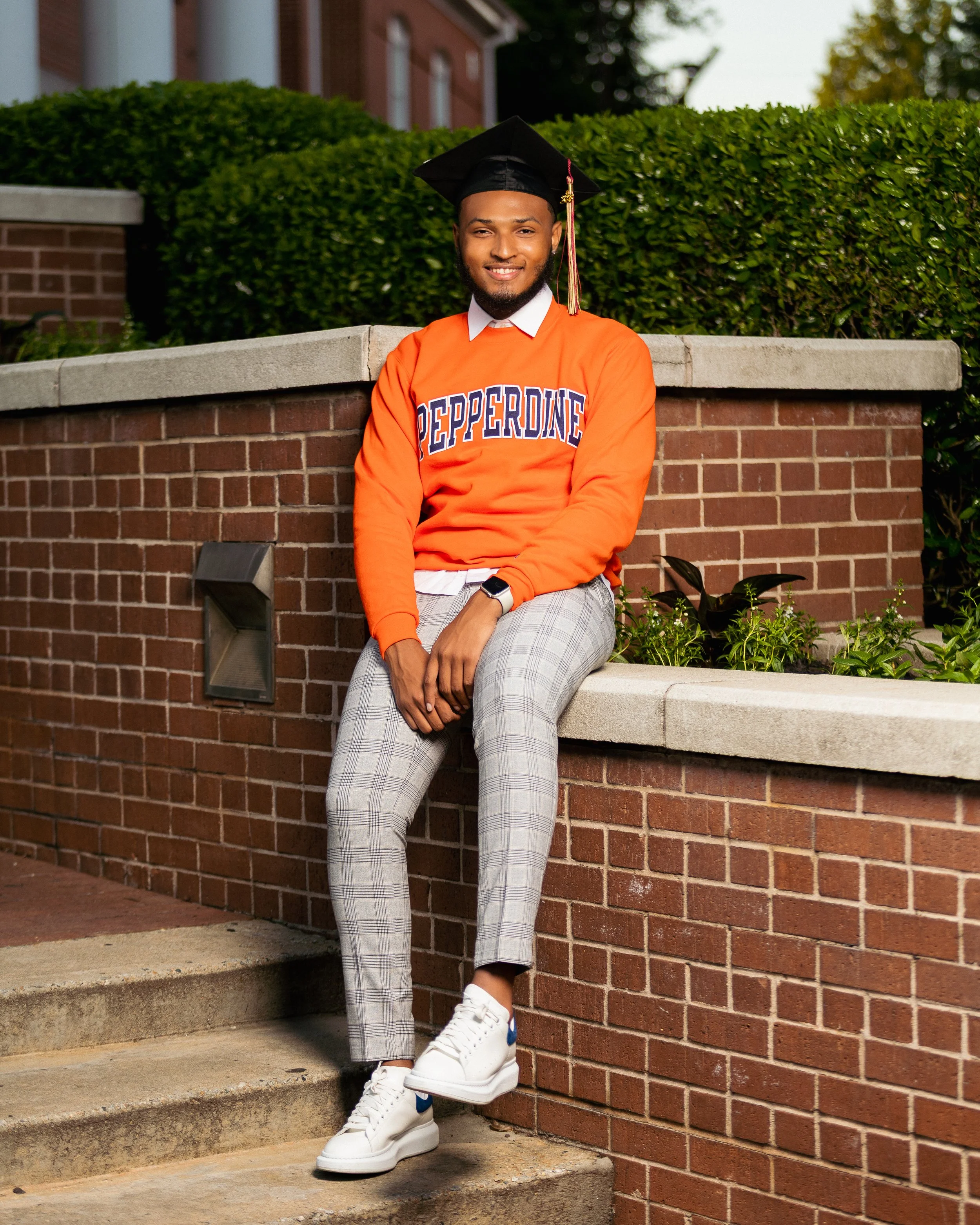 A young man sitting on a brick staircase outdoors, wearing graduation cap and an orange sweatshirt with 'Pepperdine' written on it, plaid pants, and white sneakers, smiling at the camera.