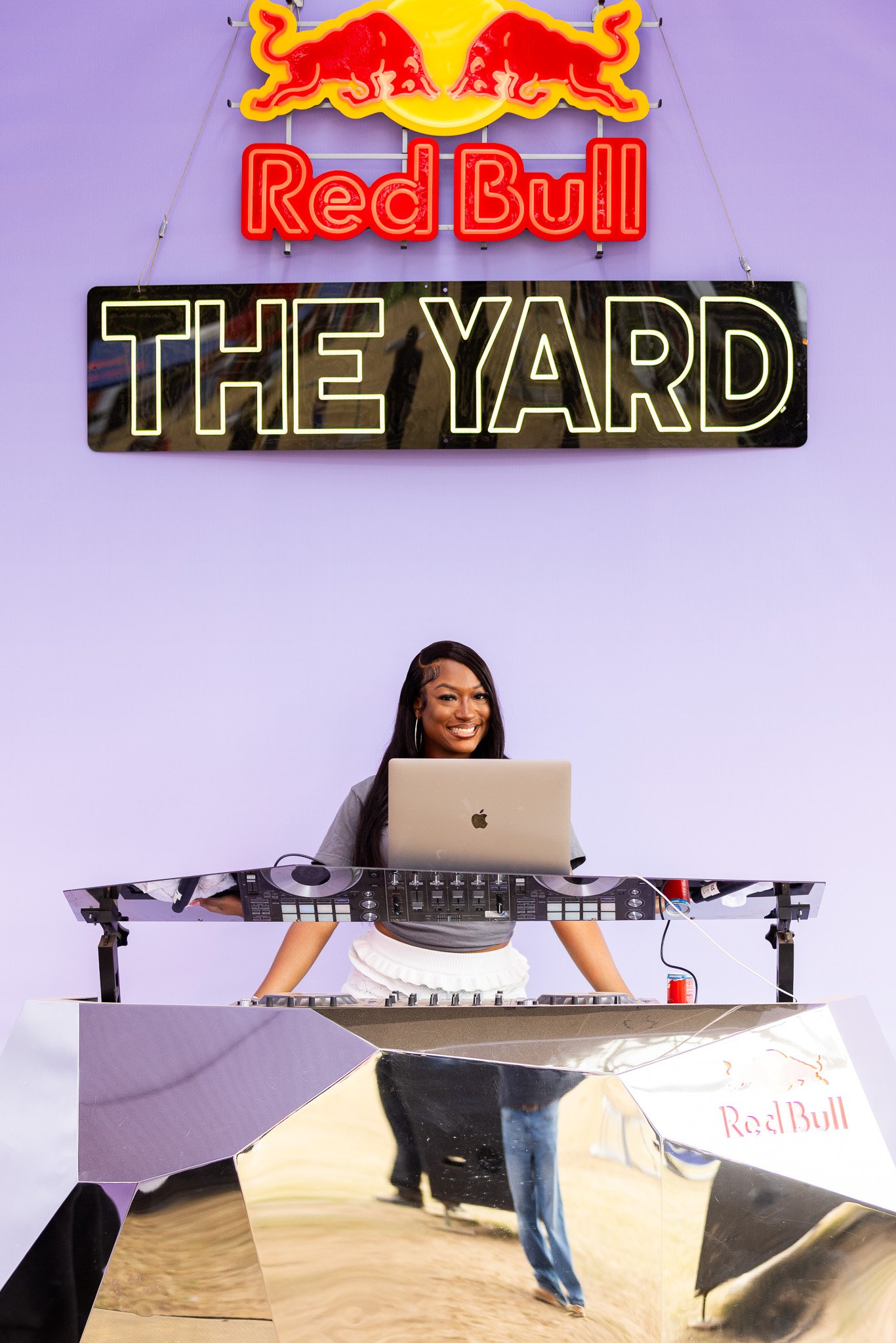 A woman is standing behind a DJ setup with a laptop and a mixer, smiling. Above her, there are illuminated signs that say 'Red Bull' and 'THE YARD', with the Red Bull logo featuring two red bulls charging at each other against a yellow sun background