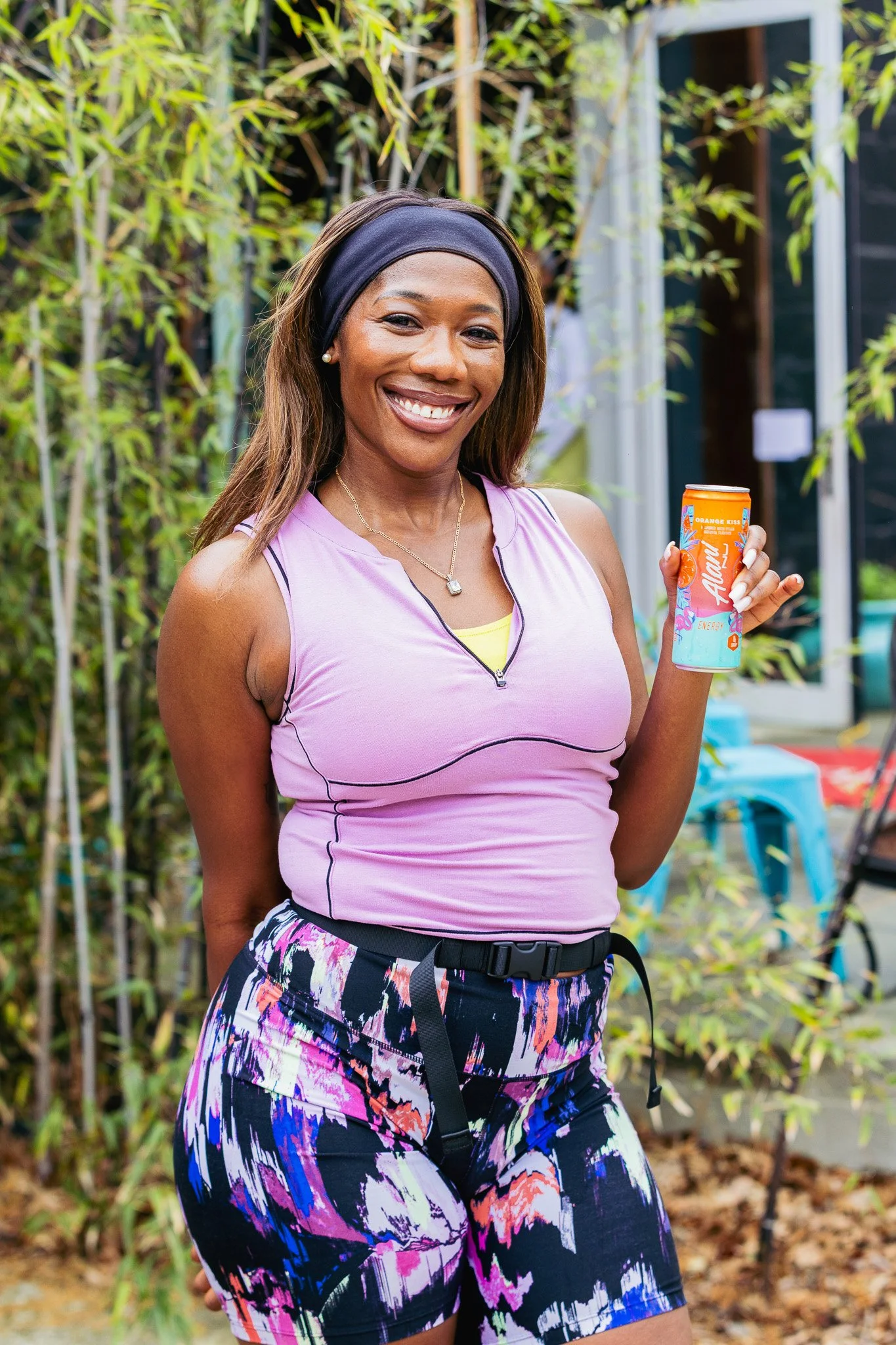 Smiling woman holding a can of drink outdoors, wearing a purple athletic top, colorful patterned shorts, and a black headband, with green plants in the background.