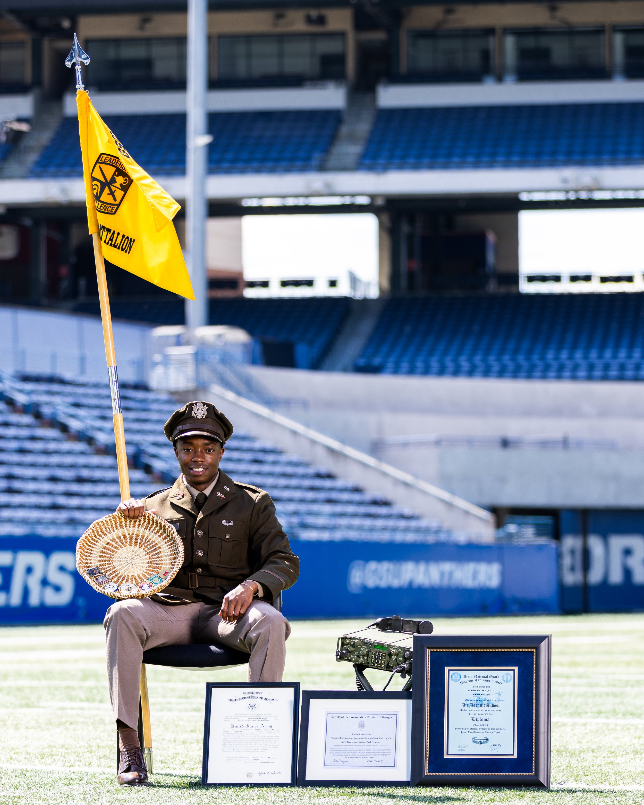 A young man dressed in military uniform sitting on a chair at a stadium, holding a yellow flag with a black emblem and text, with framed certificates and awards on display in front of him. The background shows empty stadium seats and part of the stad