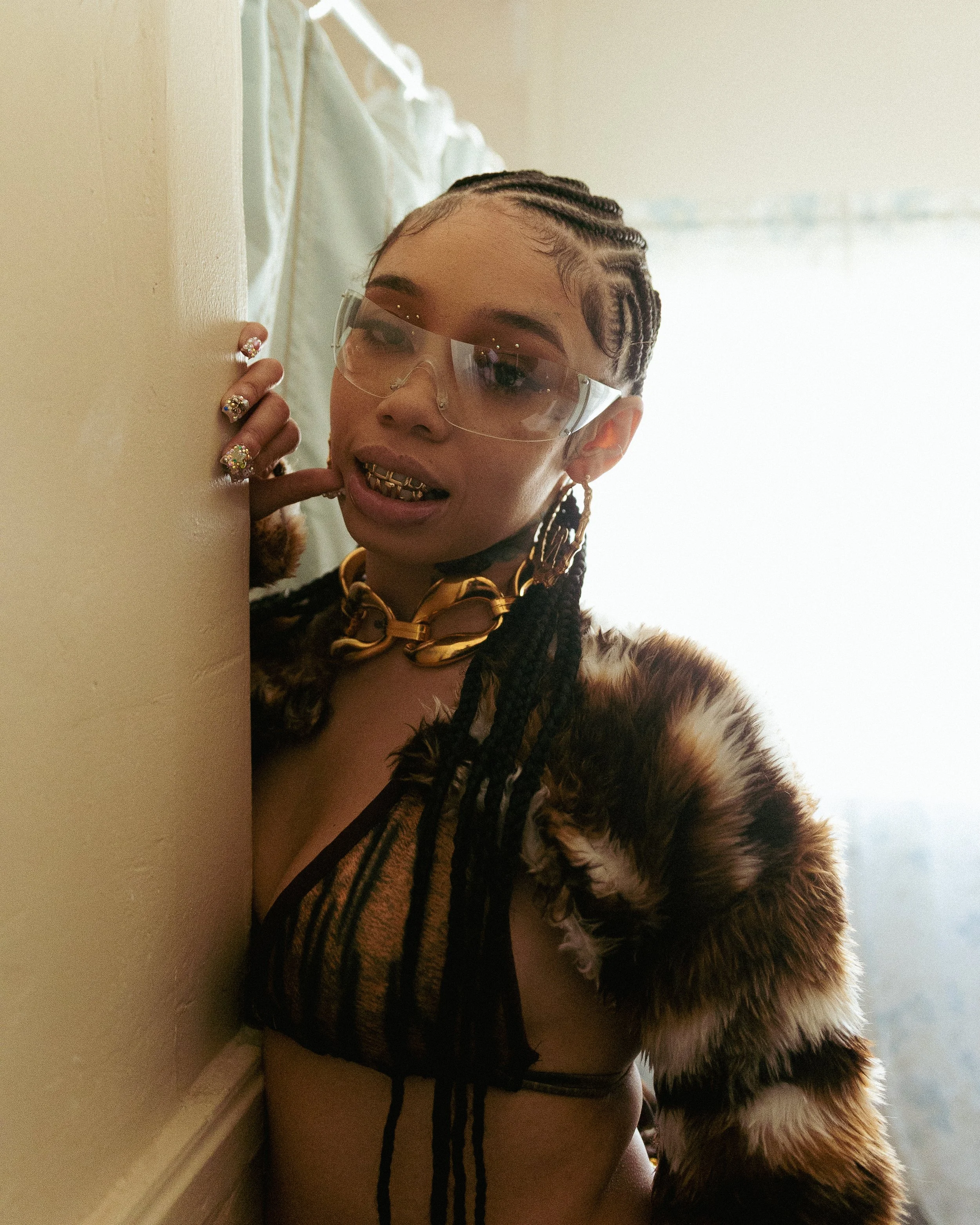A woman with braided hair wearing large earrings, a gold chain, protective glasses, a furry coat, and gold grillz, is posing near a wall with a neutral expression.