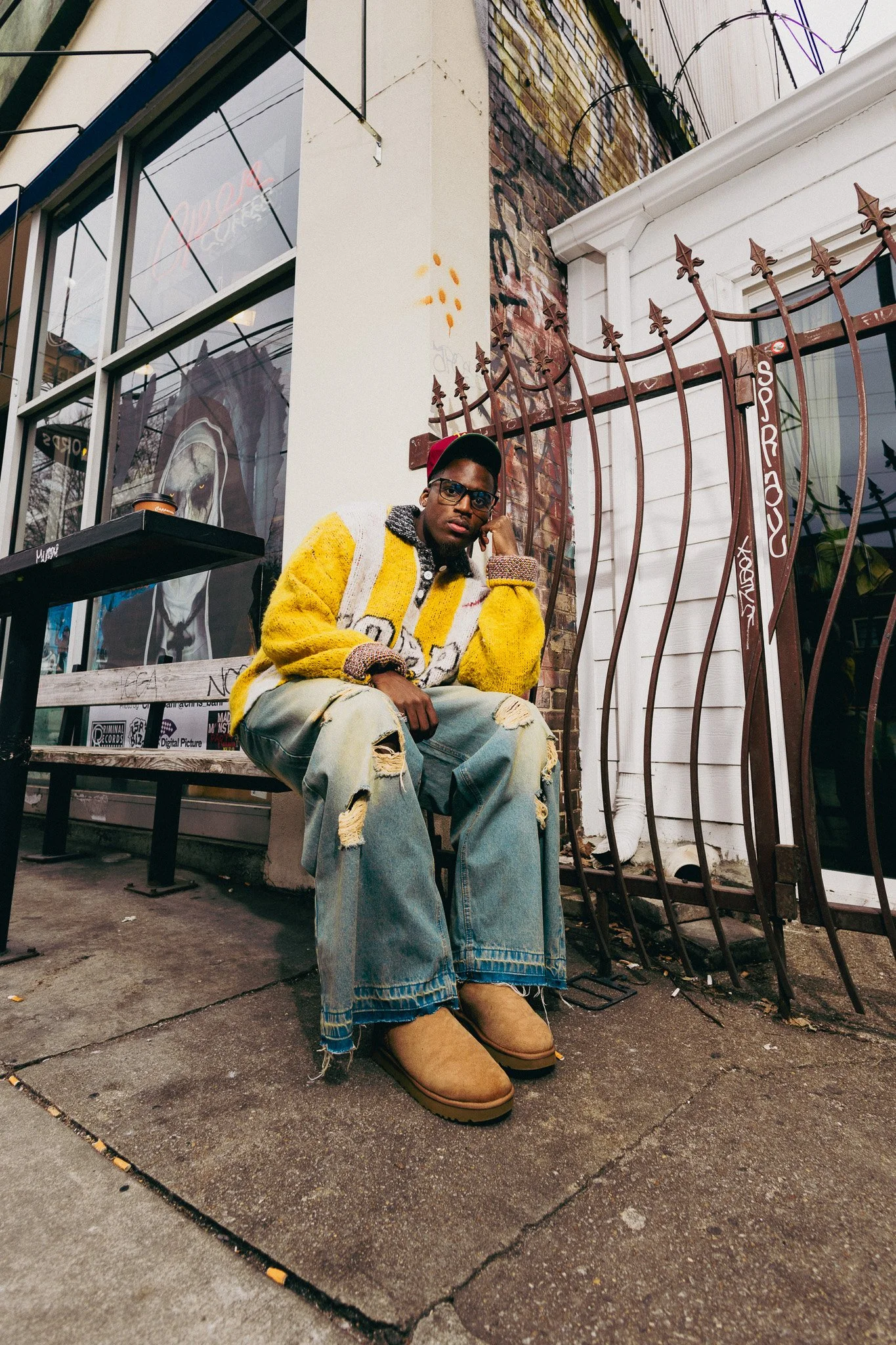 A young man sitting on a sidewalk outside a storefront with large windows. He is wearing a yellow and white sweater, ripped jeans, tan boots, and glasses. There is a graffiti and brick wall behind him, and a metal fence nearby. The window shows a lio