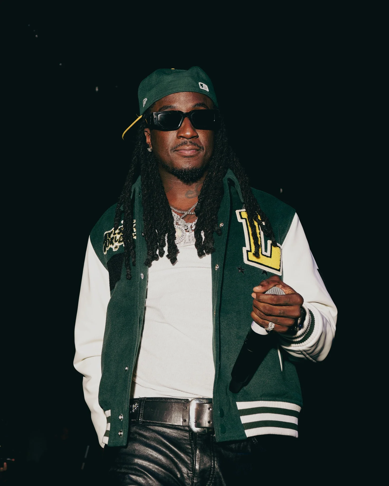 A man with long dreadlocks wearing a green baseball cap, black sunglasses, a varsity jacket with patches, a white shirt, and shiny black leather pants, holding a microphone.
