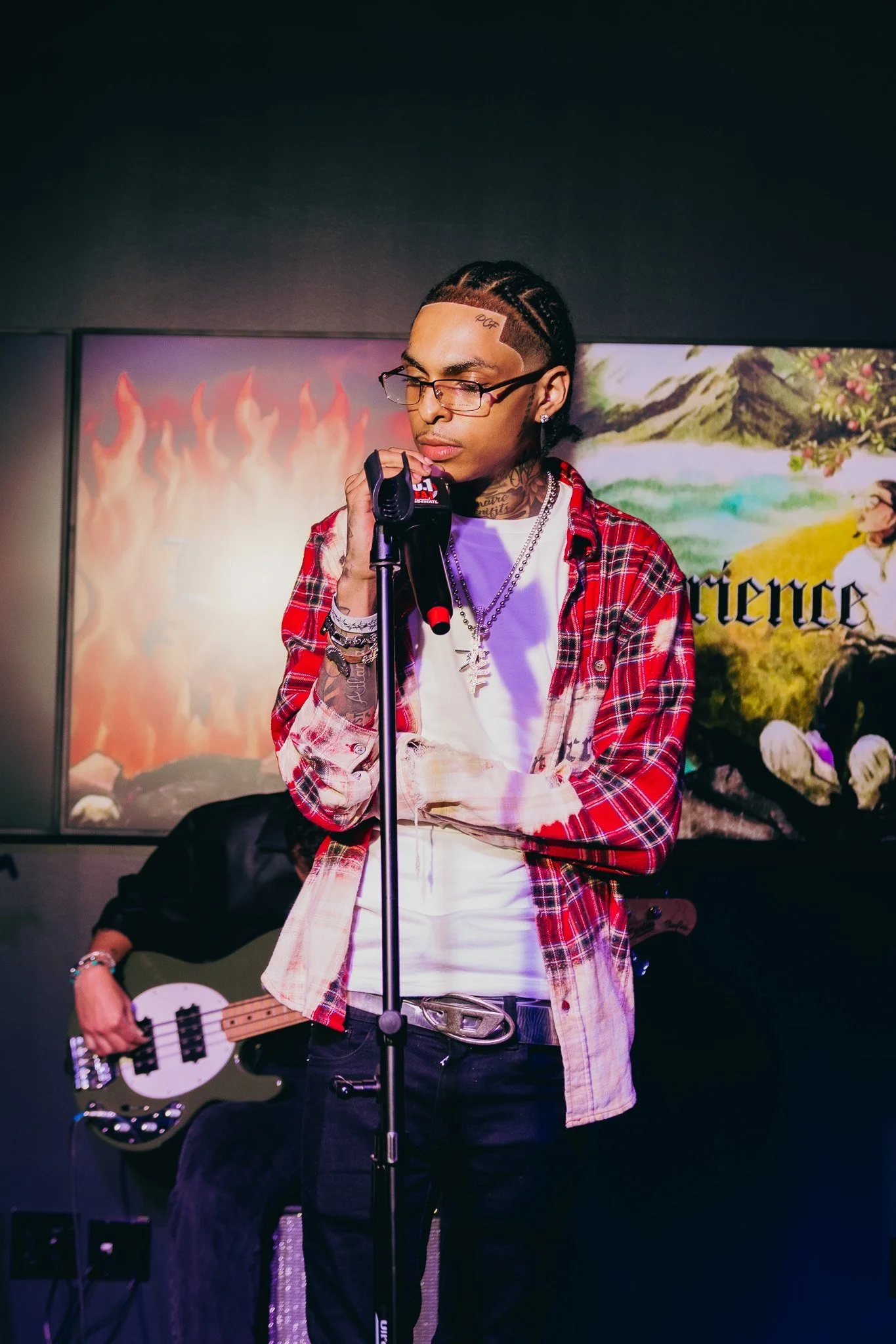 A young man with glasses and cornrow hairstyle singing into a microphone at a music performance, wearing a red plaid shirt and a white t-shirt with tattoos visible on his neck and arms, with a person playing bass guitar in the background and colorful