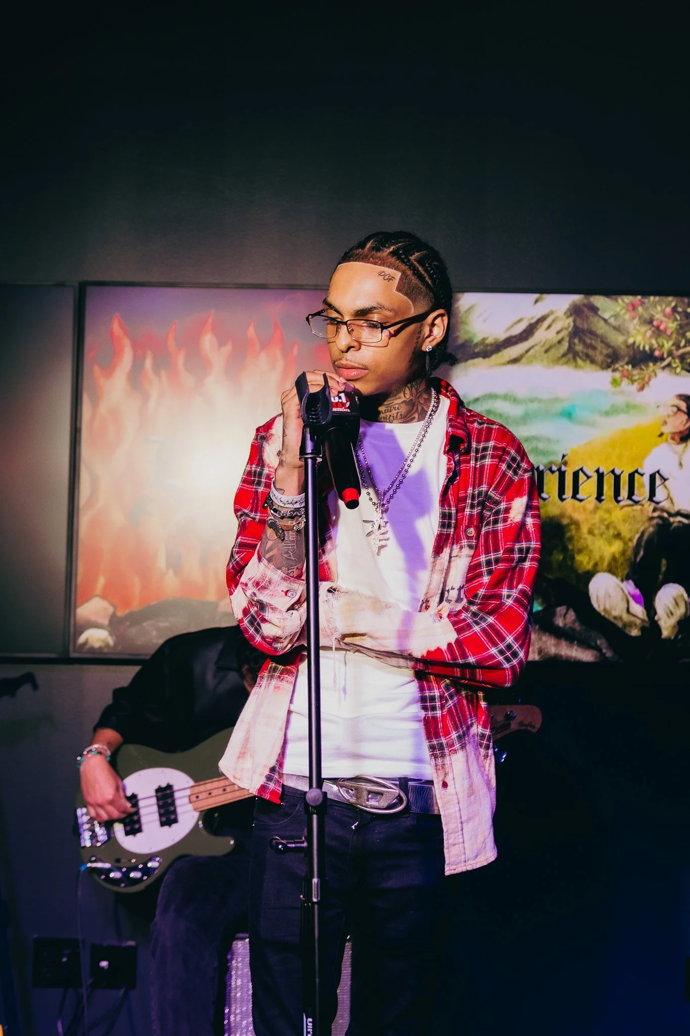 A young man with glasses, tattoos, and long hair styled in cornrows, dressed in a red plaid shirt and white t-shirt, is singing into a microphone while holding a guitar, with colorful paintings in the background.