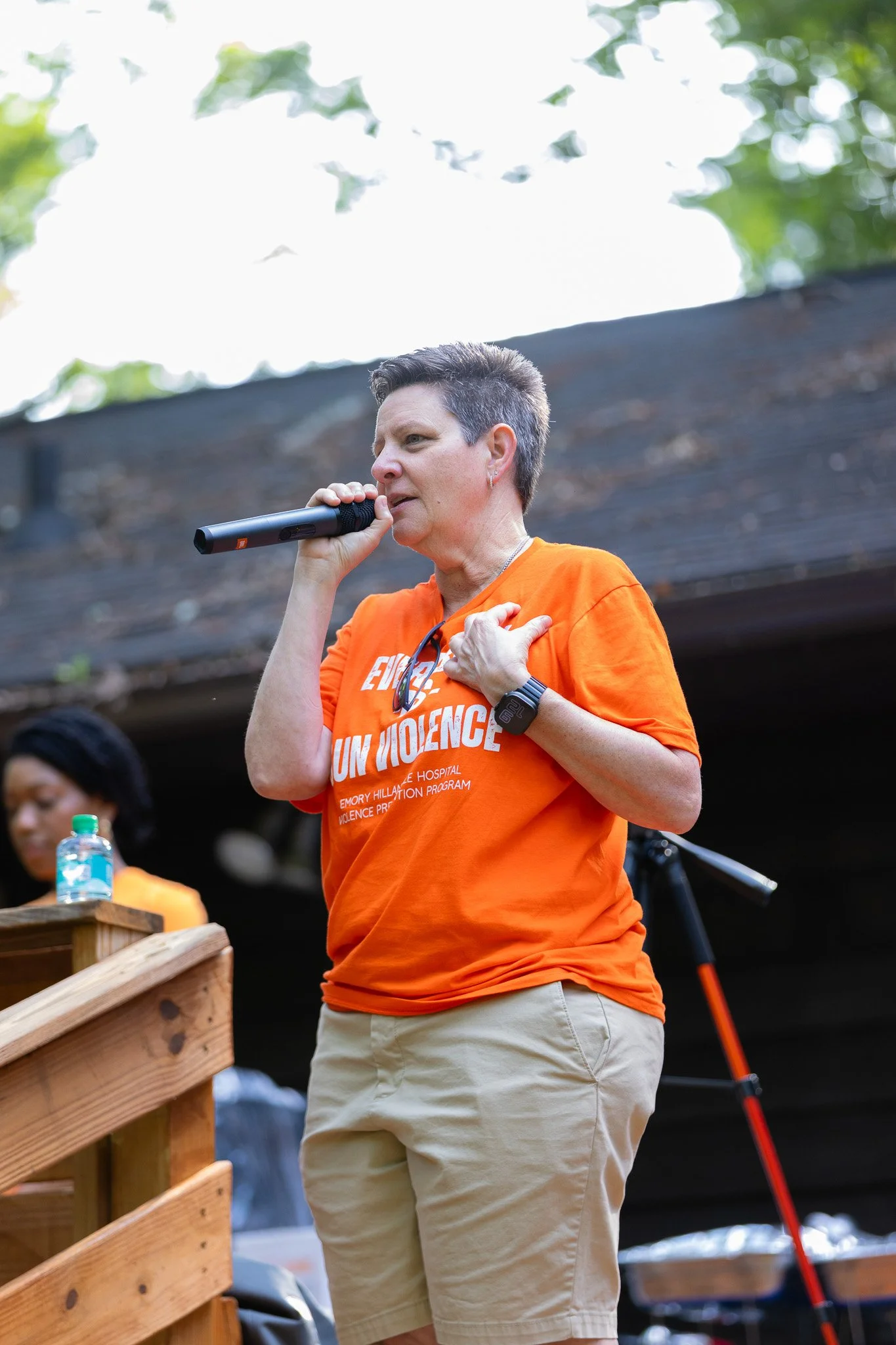 A woman with short dark hair speaking into a microphone during an outdoor event, wearing an orange T-shirt that reads 'End Violence' and beige shorts, with trees and a wooden railing in the background.