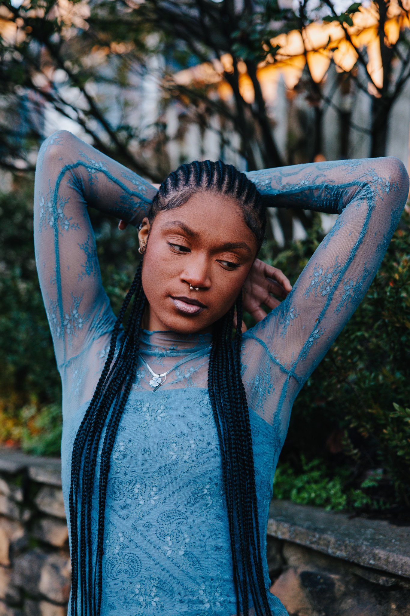 A woman with braided hair and piercings wears a sheer blue lace dress, standing outdoors with trees in the background during sunset.