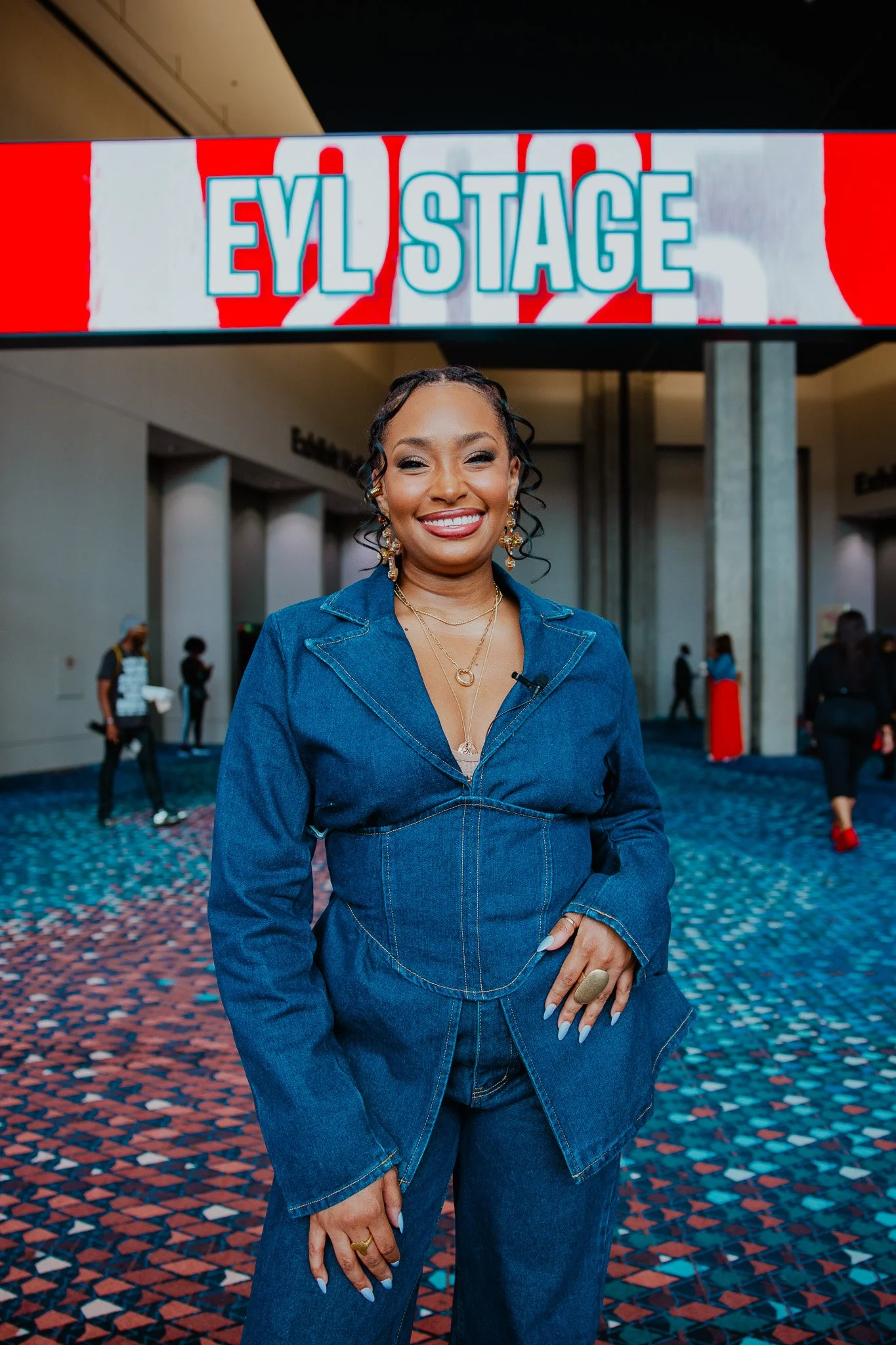 A woman with curly hair wearing a denim jacket and jeans standing in front of a sign that reads 'EYL STAGE' at an indoor event.