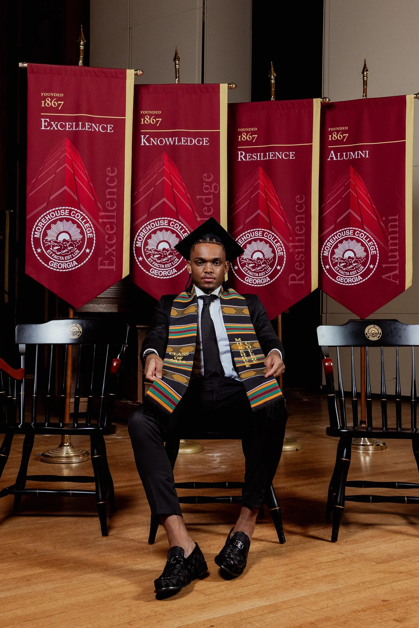 A man sitting in a chair wearing a graduation cap and gown with a colorful stole, in front of four red banners with gold trim that say 'Founded 1867,' 'Excellence,' 'Knowledge,' 'Resilience,' and 'Alumni,' at Morehouse College in Georgia.