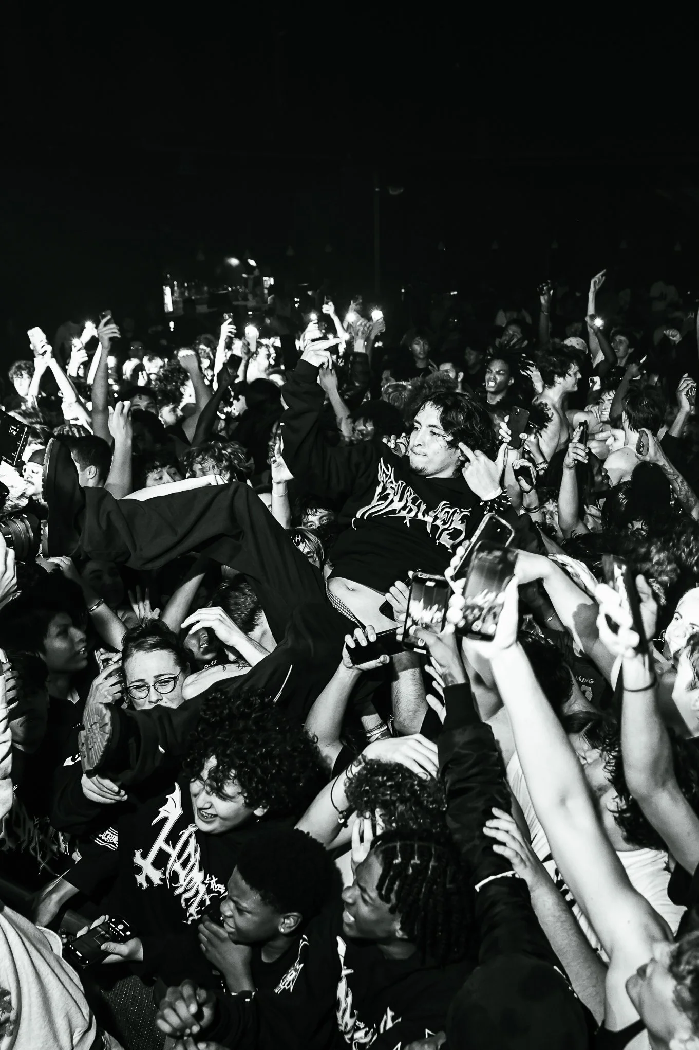 Crowd at a concert or music event, with many people raising their hands and holding phones, and an individual being lifted above the crowd.