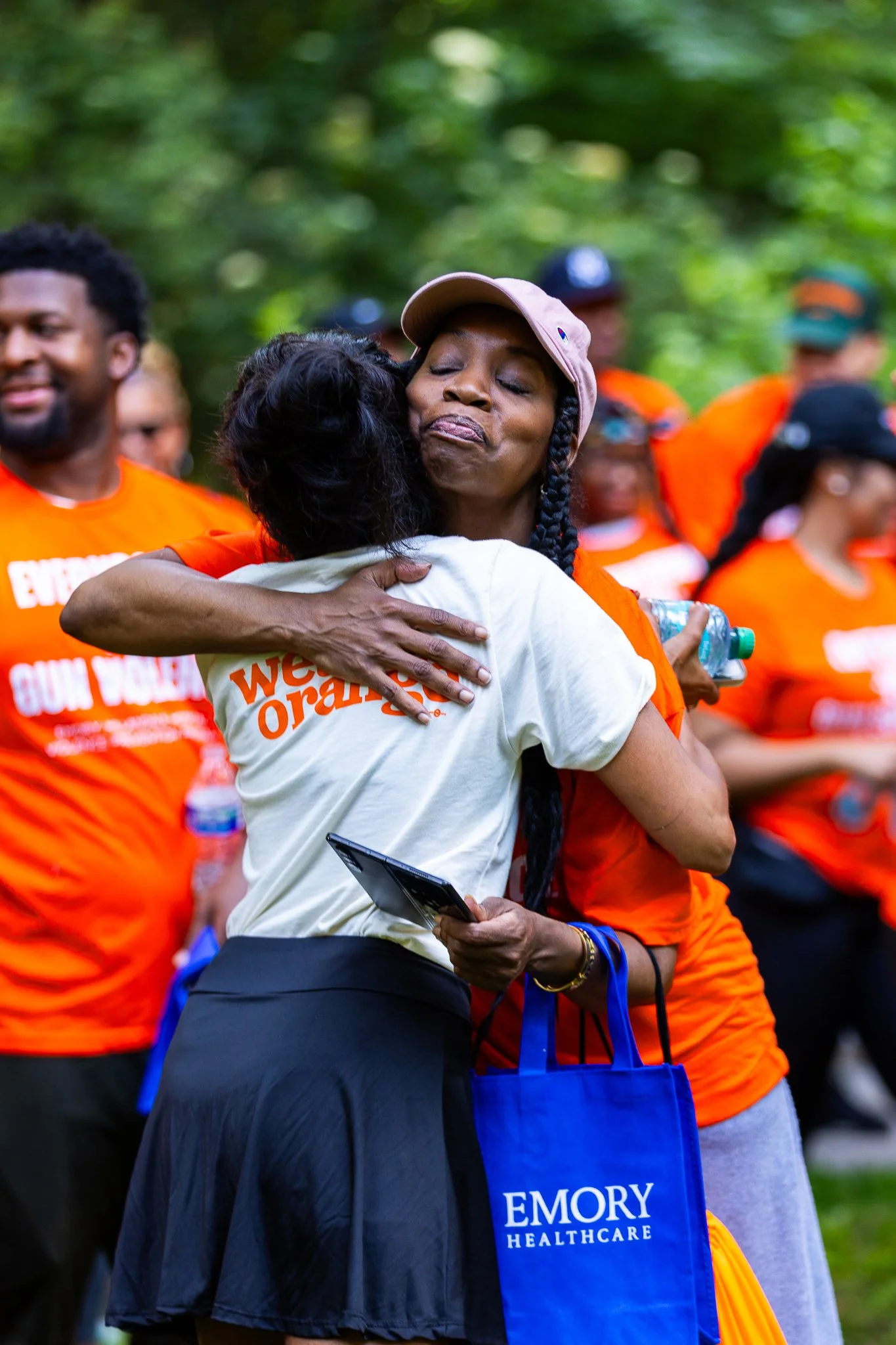 A woman wearing a pink cap and orange shirt hugging a person in a gray shirt at an outdoor gathering. Other people, some in orange shirts, are in the background.